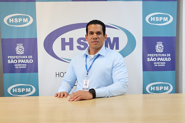 Foto colorida em ambiente interno. Um homem adulto, de pele morena, cabelo curto e escuro, está sentado atrás de uma mesa clara, de frente para a câmera, com expressão serena. Ele usa camisa social azul clara, relógio no pulso e crachá pendurado no pescoço, com as mãos apoiadas sobre a mesa. Ao fundo, há um painel institucional com repetição de logotipos e textos “HSPM” e “Prefeitura de São Paulo – Secretaria da Saúde”, em tons de azul, verde e branco. O ambiente é bem iluminado e organizado, com aparência formal e institucional.