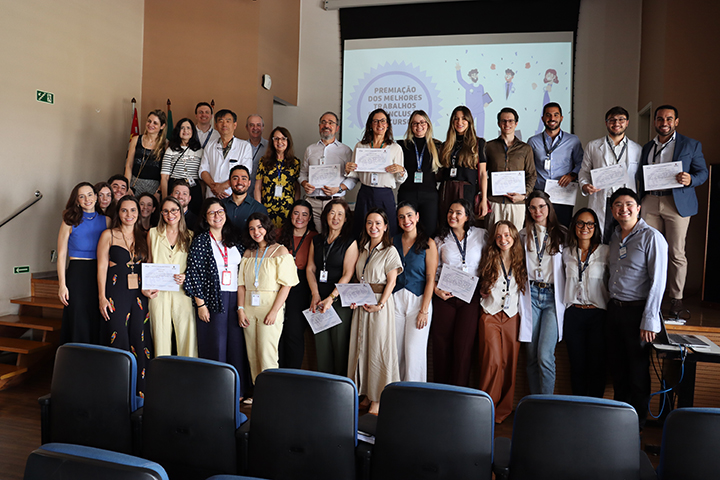 Foto colorida em um auditório. Um grande grupo de pessoas adultas está reunido no palco e à frente dele, posando para a foto e sorrindo. Homens e mulheres de diferentes idades e tons de pele seguram certificados nas mãos. As pessoas usam roupas sociais e casuais em cores variadas. Ao fundo, há um telão com uma apresentação projetada, com ilustrações de pessoas e texto indicando um evento de premiação ou reconhecimento. As cadeiras do auditório aparecem em primeiro plano, vazias. O ambiente é bem iluminado e transmite um clima de celebração, conquista e reconhecimento coletivo.