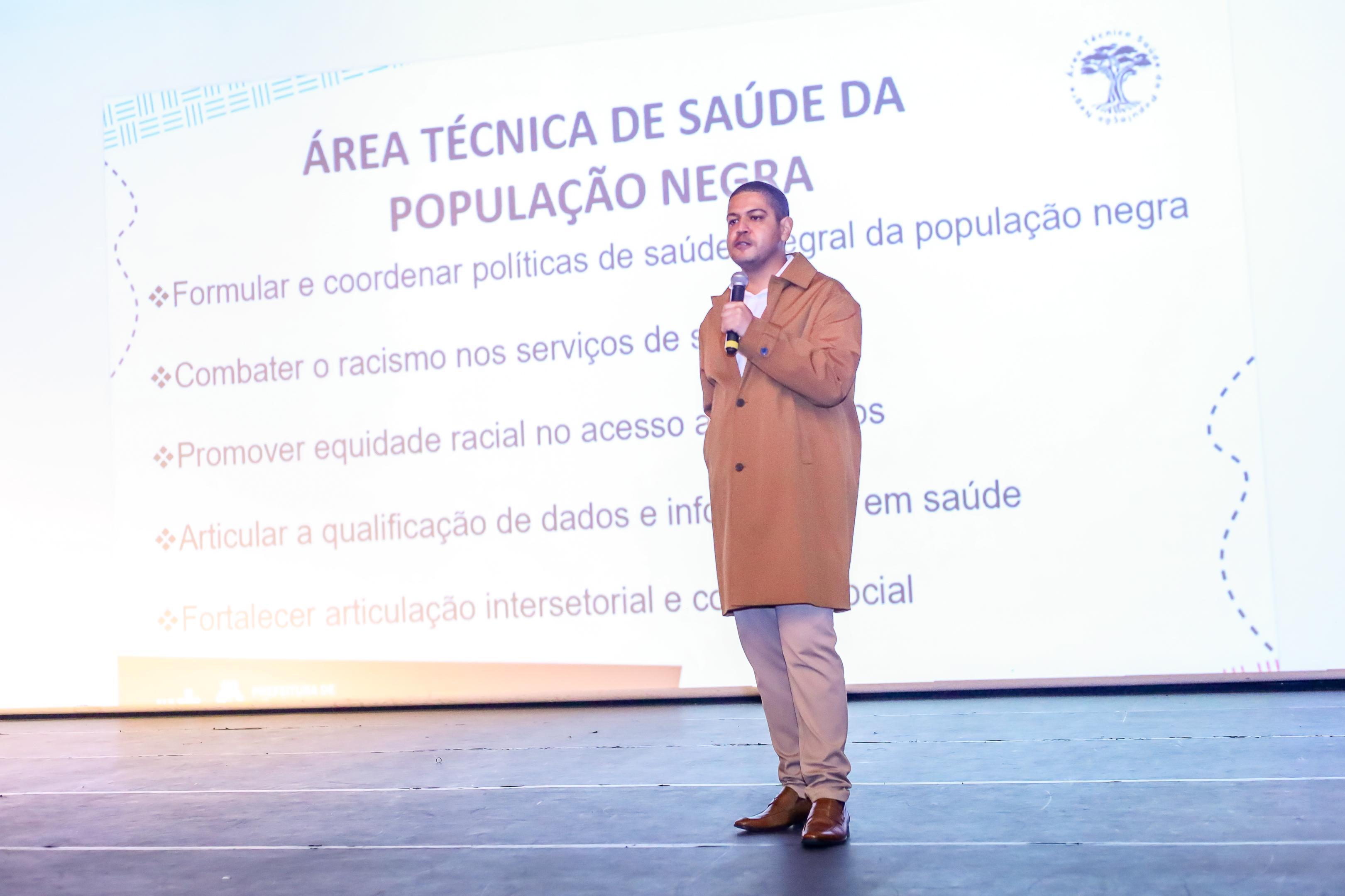 A imagem mostra Daniel, que está em pé, segurando um microfone e falando diante de uma grande tela de projeção em um auditório. Ele tem pele clara, cabelo curto e escuro, e veste um casaco marrom claro que vai até os joelhos, calça bege e sapatos marrons. Atrás dele, o telão exibe o título “Área Técnica de Saúde da População Negra”, seguido de um texto que descreve as funções desse setor, como a formulação e coordenação de políticas de saúde integral da população negra, o combate ao racismo nos serviços de saúde, a promoção da equidade racial no acesso aos atendimentos e a qualificação de dados e informações em saúde.