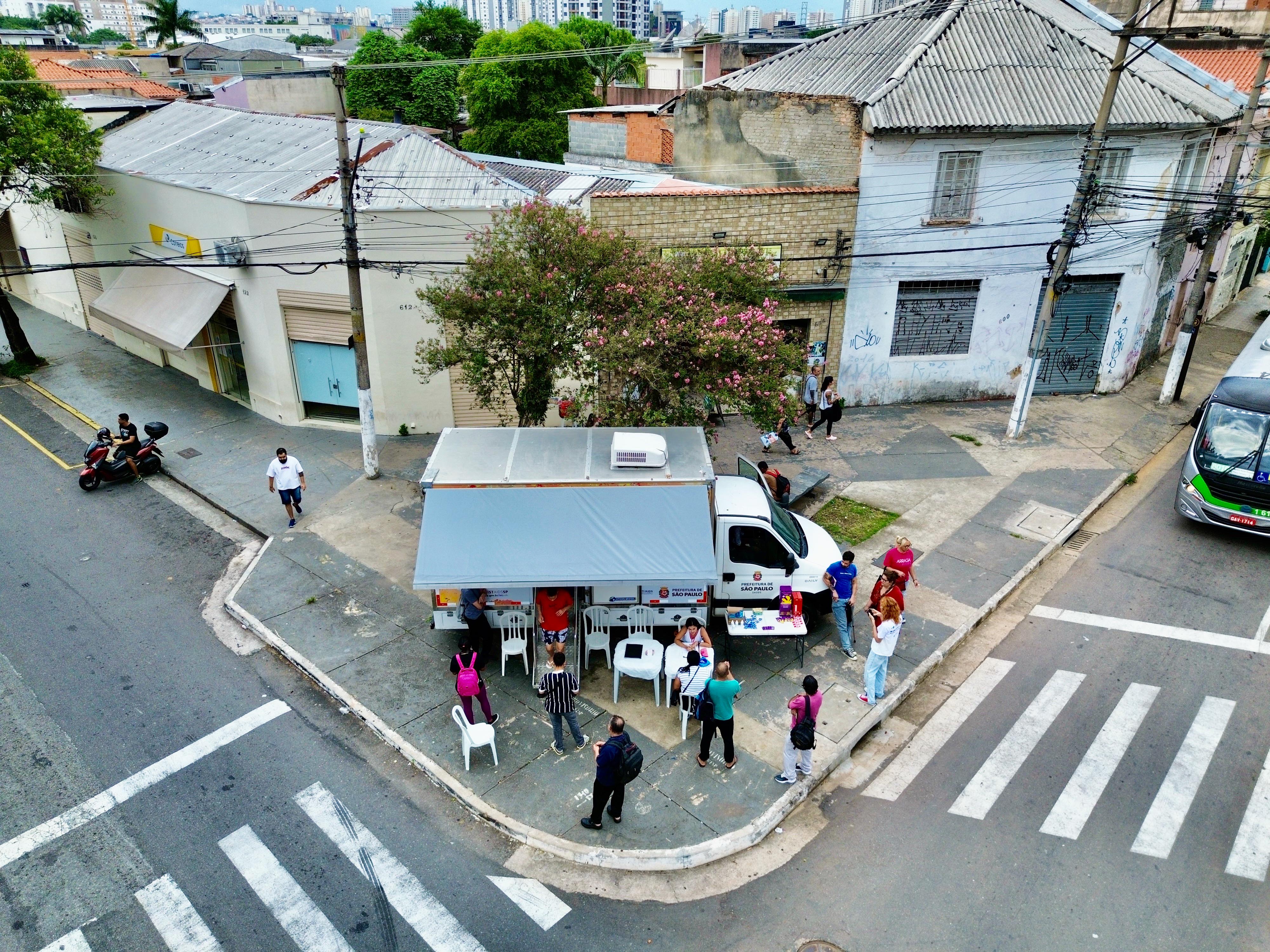 A imagem mostra uma cena urbana registrada de cima, em uma esquina de bairro, provavelmente em uma região central ou mista da cidade. No centro da foto, há um veículo adaptado da Prefeitura de São Paulo estacionado na calçada, funcionando como uma unidade móvel de atendimento. Ele possui uma cobertura lateral estendida, sob a qual estão dispostas cadeiras e uma pequena mesa, formando um espaço de acolhimento para o público. Em frente e ao redor do veículo, várias pessoas aguardam atendimento ou conversam com profissionais, formando uma pequena fila. Algumas estão sentadas, enquanto outras permanecem em pé. A movimentação sugere a oferta de algum serviço público, possivelmente relacionado à saúde ou assistência social. O local é uma esquina com faixas de pedestres bem demarcadas e calçadas amplas. Ao fundo, aparecem casas e prédios antigos, com fachadas desgastadas, muros grafitados e telhados de diferentes materiais. Uma árvore com flores rosadas se destaca no meio da paisagem, trazendo contraste com o concreto ao redor