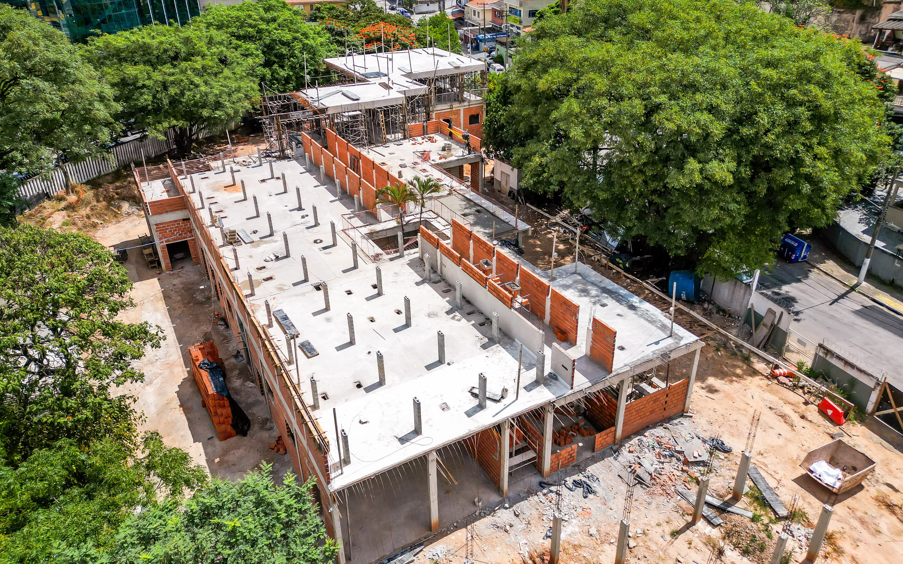 A imagem mostra uma obra em andamento vista de cima, provavelmente registrada por drone. Trata-se de um prédio de médio porte em construção, com estrutura de concreto já concluída em parte e paredes de tijolos aparentes sendo erguidas.  É possível ver lajes prontas com diversos pilares verticais expostos, indicando que o edifício terá mais de um pavimento. Em algumas áreas, há andaimes montados e trabalhadores atuando, sugerindo que a obra está em fase estrutural e de alvenaria.  O entorno é bastante arborizado, com árvores grandes ocupando boa parte do terreno ao lado e criando contraste com a construção. Também há uma rua próxima, com calçada, postes e alguns equipamentos urbanos visíveis.  No chão, ao redor da obra, aparecem materiais de construção espalhados, como tijolos, entulho e caçambas, além de áreas de terra batida.