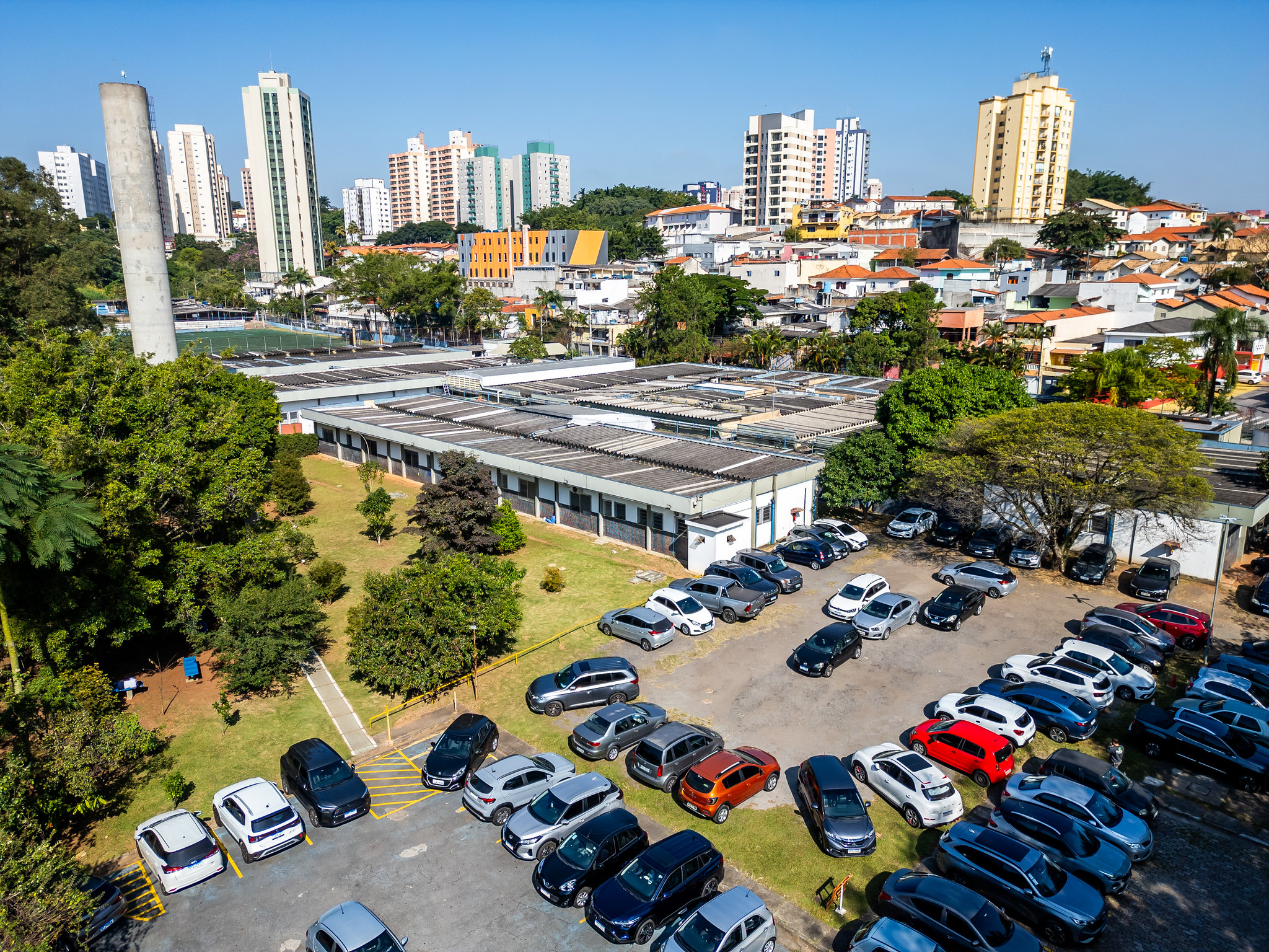Vista aérea do Hospital Municipal e Maternidade Professor Mário Degni, um importante centro de saúde pública localizado na Zona Oeste de São Paulo. A foto captura o contraste entre as instalações horizontais do hospital e o denso cenário urbano ao fundo. É possível ver dezenas de veículos de diversas cores (branco, prata, preto, azul, vermelho e até um laranja vibrante). Alguns carros estão estacionados em vagas demarcadas no asfalto, enquanto outros ocupam áreas que parecem ser de terra ou grama. Ao fundo, a linha do horizonte é dominada por vários edifícios residenciais altos de cores claras (branco e bege), típicos da região do Rio Pequeno/Butantã.