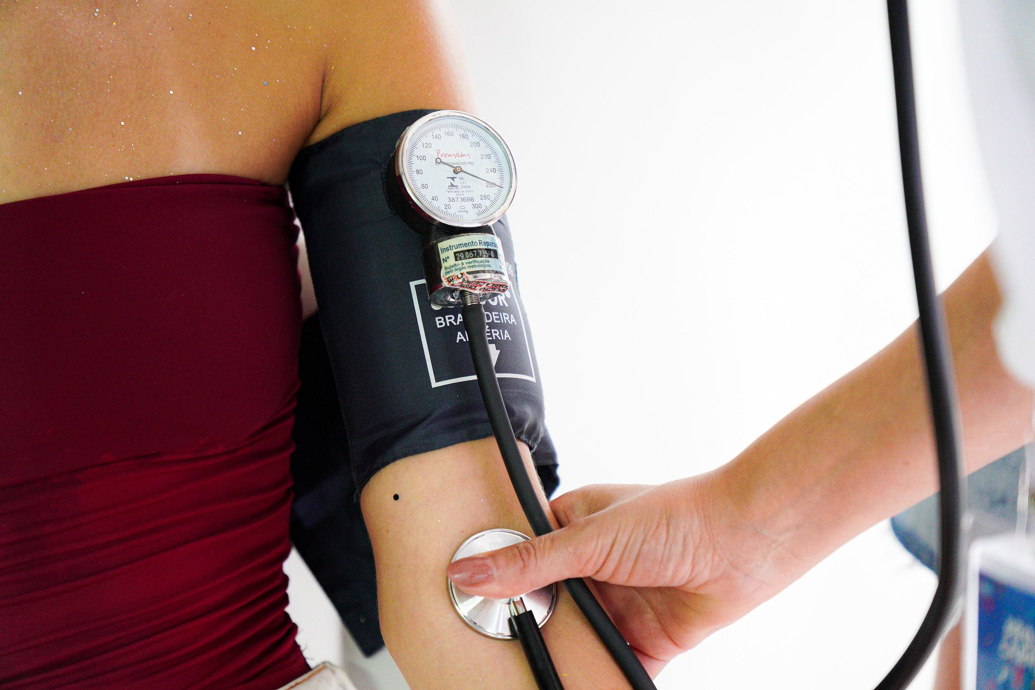 A imagem mostra um atendimento de saúde em andamento, com foco no cuidado e na prevenção durante o pré-Carnaval.  Em primeiro plano, vemos o braço de uma pessoa sendo aferido com um aparelho de pressão arterial. O manguito está ajustado ao braço, ligado a um medidor analógico, enquanto a profissional de saúde utiliza o estetoscópio para ouvir os batimentos e realizar a leitura. O enquadramento é fechado, destacando o gesto técnico e a atenção ao paciente.