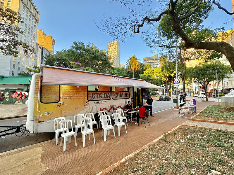 A imagem mostra uma unidade móvel de atendimento instalada em uma praça ou área pública arborizada de uma região central da cidade. Em destaque está um trailer adaptado, identificado como “CTA da Cidade”, sigla normalmente associada aos Centros de Testagem e Aconselhamento em saúde. O veículo possui uma cobertura retrátil aberta sobre a calçada, criando uma área de atendimento ao público. Em frente ao trailer há várias cadeiras plásticas brancas organizadas para espera, enquanto algumas pessoas conversam ou realizam atendimentos em mesas instaladas ao lado da unidade. O cenário urbano é composto por prédios altos ao fundo, árvores de grande porte, palmeiras e uma praça com áreas de circulação de pedestres. A luz do sol ilumina a cena, sugerindo que a atividade ocorre durante o dia.