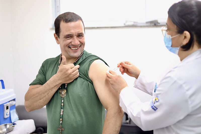 A imagem mostra uma cena de vacinação em um ambiente de saúde. Um homem adulto, vestindo uma camiseta verde e um colar com contas e um crucifixo, sorri enquanto recebe uma injeção no braço esquerdo. Com uma das mãos, ele segura a manga da camiseta levantada para facilitar a aplicação. Sua expressão é descontraída e positiva, transmitindo confiança e tranquilidade durante o procedimento. À direita da imagem, uma profissional de saúde, vestida com jaleco branco e máscara cirúrgica azul, prepara ou aplica a vacina. Ela aparece de perfil, concentrada no atendimento. O ambiente é limpo, bem iluminado e tem características típicas de um consultório ou sala de vacinação, com equipamentos médicos e superfícies clínicas ao fundo.
