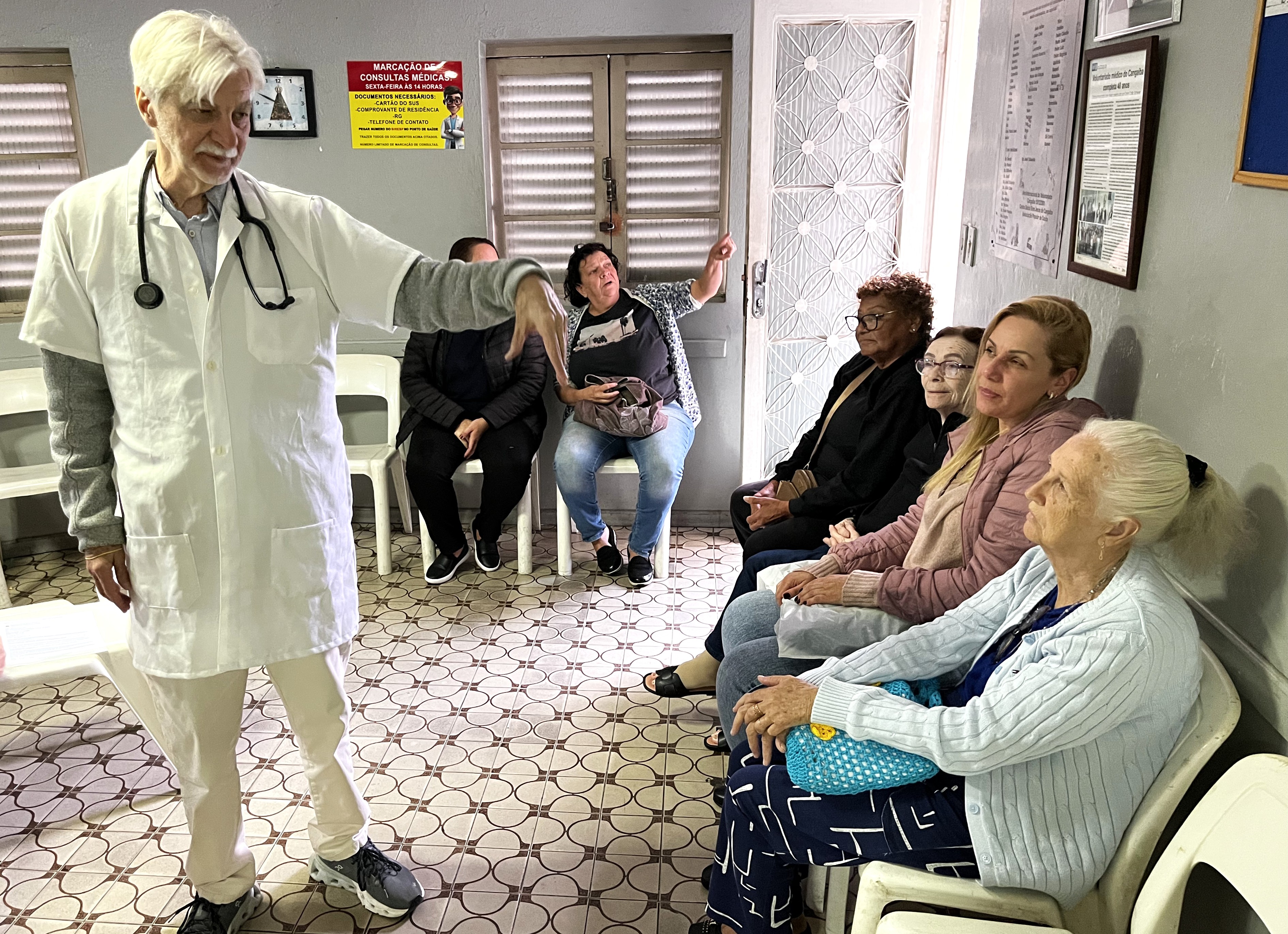 A imagem mostra uma atividade de educação em saúde realizada em uma sala de espera de uma unidade de saúde.  Em primeiro plano, um profissional de saúde, vestindo jaleco branco e com um estetoscópio pendurado no pescoço, conversa com um grupo de pacientes. Ele faz um gesto com a mão enquanto parece explicar algum tema ou orientar os presentes.  Sentadas em cadeiras ao longo da parede, estão principalmente mulheres, incluindo pessoas idosas, que acompanham atentamente a atividade. O ambiente é simples e acolhedor, com cartazes informativos, quadro de avisos, relógio e sinalizações relacionadas ao funcionamento do serviço de saúde.  A cena transmite a ideia de cuidado próximo e humanizado, destacando o papel da Atenção Primária na promoção da saúde, na orientação dos usuários e na construção de vínculos entre profissionais e comunidade. A presença de pessoas idosas sugere que a atividade pode estar relacionada a temas como envelhecimento saudável, prevenção de doenças, autocuidado ou acompanhamento de condições crônicas.