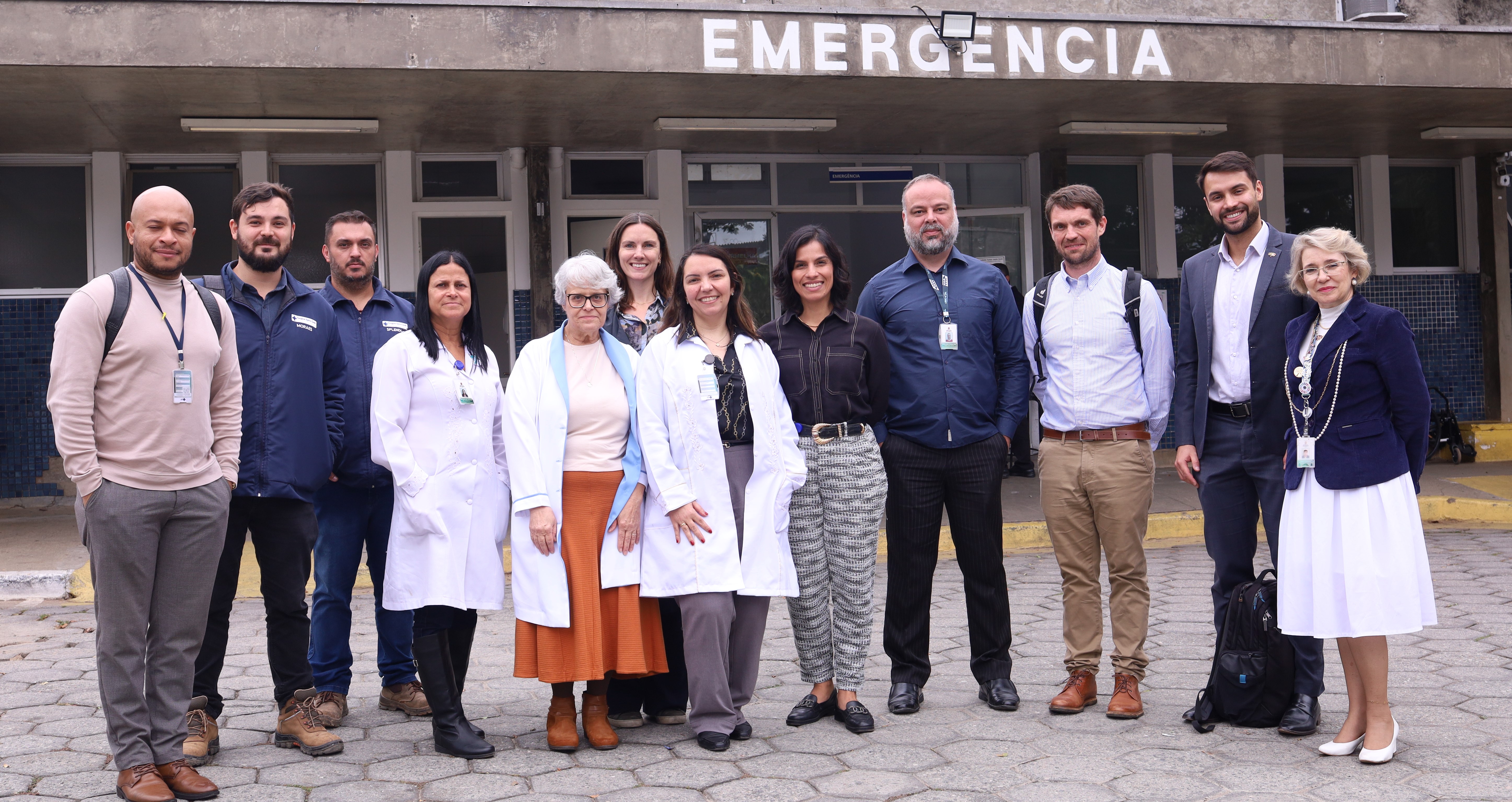 A imagem mostra um grupo de 12 pessoas em frente a uma entrada hospitalar identificada com a palavra “EMERGÊNCIA” em letras grandes na parte superior da fachada.  O grupo é composto por homens e mulheres, alguns vestidos com jalecos brancos, sugerindo que são profissionais de saúde, enquanto outros usam roupas sociais ou casuais, incluindo crachás de identificação.  A cena transmite a ideia de uma visita institucional ou registro oficial, reunindo diferentes profissionais da área da saúde e possivelmente gestores, em frente ao setor de emergência de um hospital.