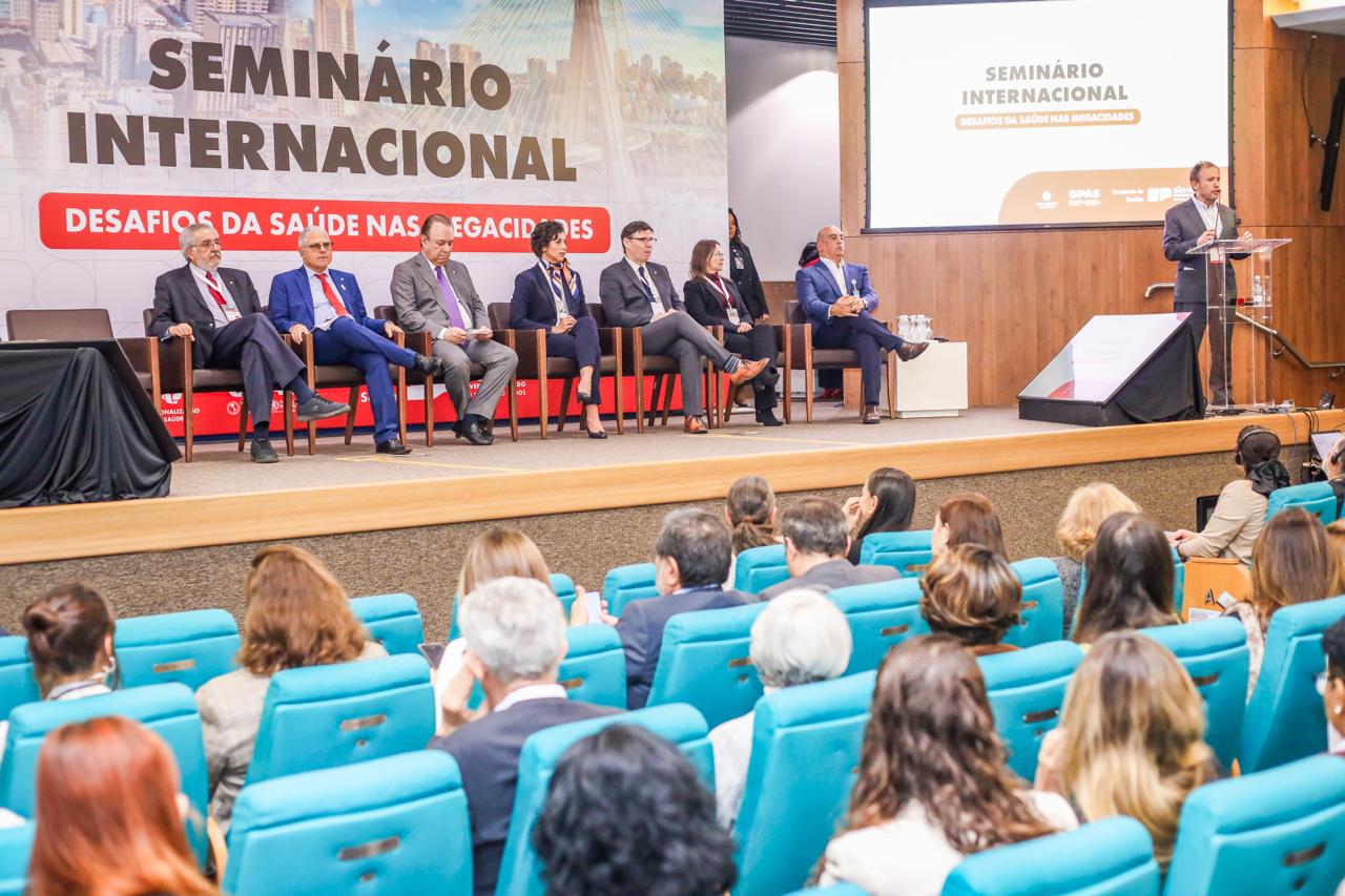 A imagem mostra um auditório durante um evento formal. No palco, há um painel de oito pessoas sentadas em cadeiras, todas vestindo trajes sociais, e à direita um homem em pé falando em um púlpito transparente, também de terno e gravata.  No fundo do palco, há um grande painel com o título em destaque:  “SEMINÁRIO INTERNACIONAL – Desafios da Saúde nas Megacidades”  Ao lado, um telão projetado reforça o mesmo título do evento.  Na plateia, várias pessoas estão sentadas em cadeiras azuis, acompanhando a apresentação. O ambiente é claro, organizado e transmite a formalidade de uma conferência ou encontro técnico de alto nível.