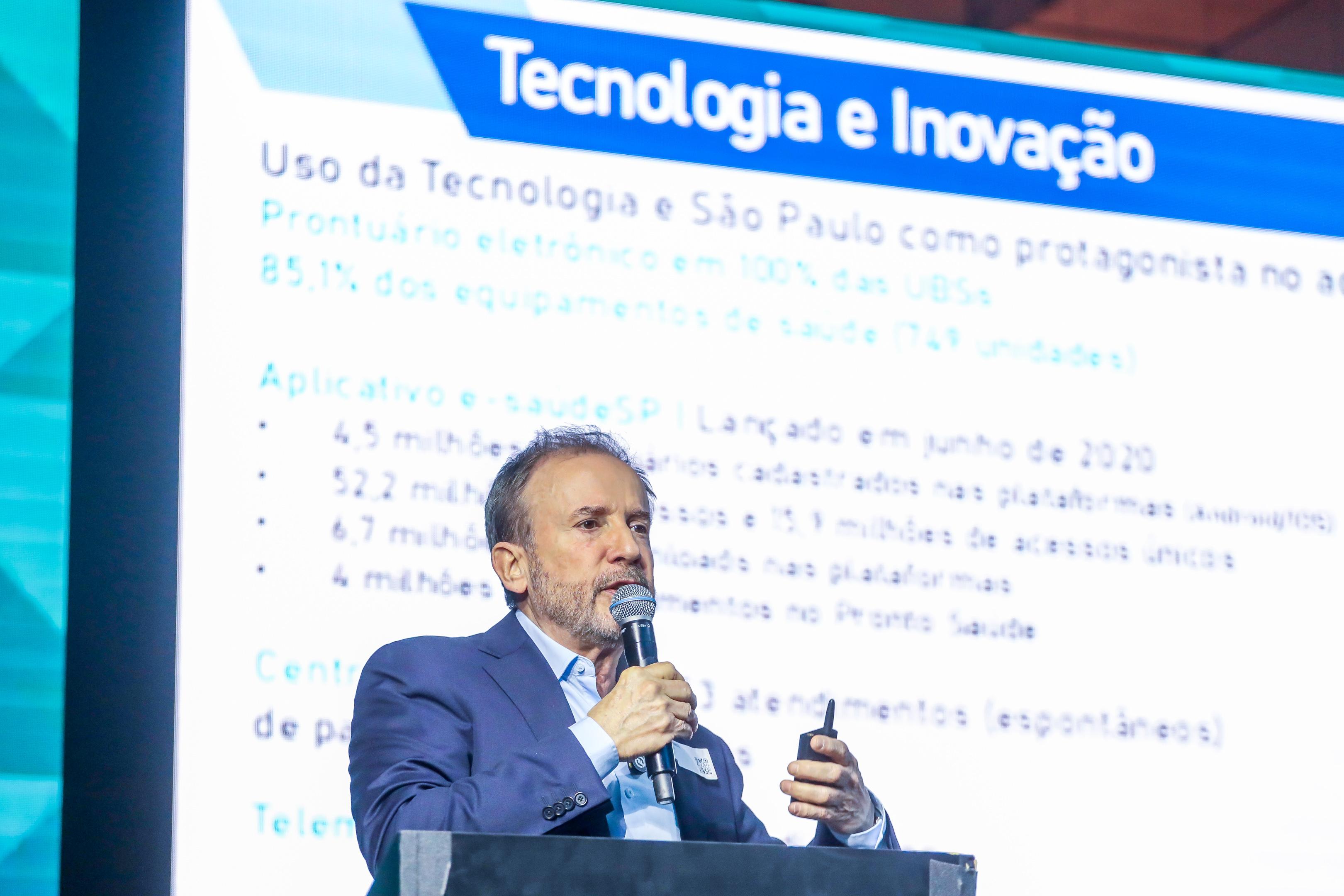 A imagem mostra O secretário da saúde, Luiz Carlos Zamarco, um homem de pele clara, com barba grisalha e cabelos curtos também grisalhos, vestido com terno azul-escuro e camisa clara. Ele segura um microfone em uma das mãos e um controle pequeno na outra, parecendo estar em uma apresentação ou palestra. Ao fundo, há uma grande tela projetada com o título em destaque “Tecnologia e Inovação” em uma faixa azul. O texto projetado fala sobre o uso da tecnologia na saúde de São Paulo, mencionando prontuário eletrônico em todas as UBSs, porcentagem de equipamentos de saúde informatizados, além de dados sobre o aplicativo e-saúdeSP, como milhões de cadastros, acessos e atendimentos.