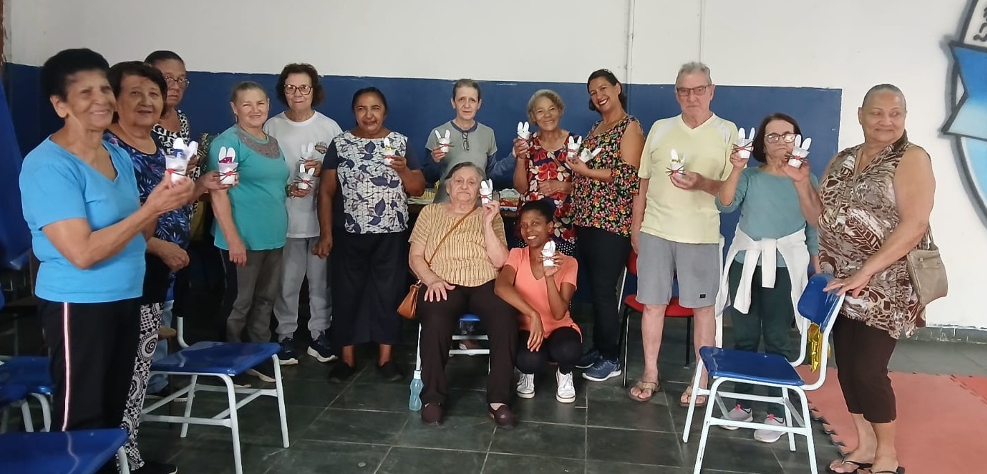 A foto mostra um grupo de aproximadamente 15 pessoas, a maioria idosas, reunidas em um espaço interno de paredes brancas e azul-marinho. Estão em pé, lado a lado, formando uma fileira, enquanto duas mulheres aparecem sentadas na frente, uma delas em uma cadeira de rodas. Todos parecem sorridentes e alegres. Eles seguram pequenas lembrancinhas em formato de coelho branco, provavelmente feitas de papel ou tecido, com detalhes coloridos, sugerindo uma atividade de artesanato ou celebração, talvez ligada à Páscoa. As pessoas vestem roupas casuais e confortáveis: camisetas, calças e blusas leves. Há diversidade nos estilos — desde estampas florais até roupas lisas. O ambiente tem cadeiras azuis de plástico e metal, algumas organizadas e outras fora de lugar, o que dá a sensação de um encontro descontraído e comunitário.