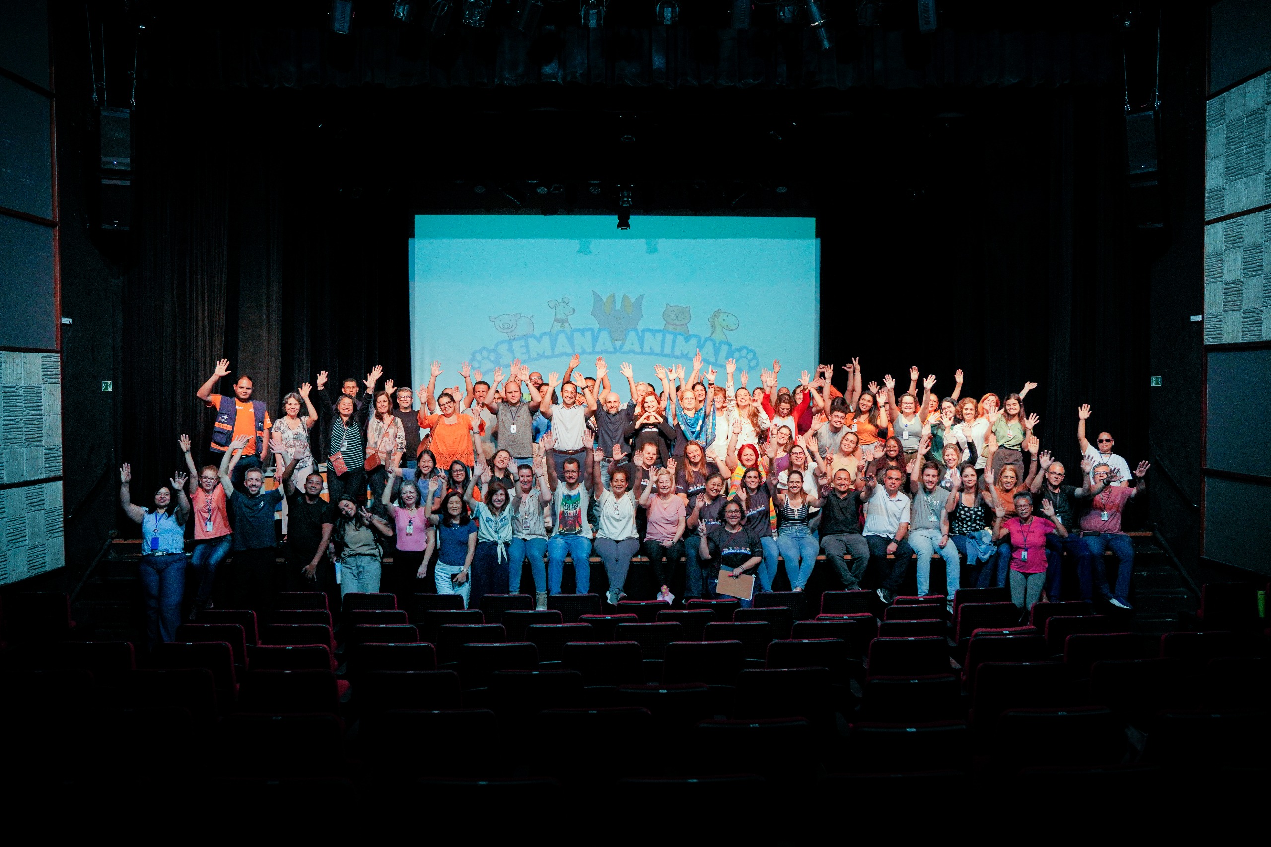 A foto mostra um grande grupo de pessoas reunidas em um teatro. Elas estão posicionadas no palco, em duas fileiras, e muitas levantam os braços em gesto de celebração e alegria. O auditório está escuro, com as poltronas vazias visíveis em primeiro plano. No fundo, há uma tela de projeção com ilustrações de animais sorridentes e a frase “Semana Animal” em letras azuis, reforçando o clima festivo e o tema do encontro. As pessoas no palco são diversas em idade, gênero e aparência, vestindo roupas casuais e coloridas. O ambiente transmite união, engajamento e espírito de confraternização, como se fosse o encerramento de um evento coletivo.