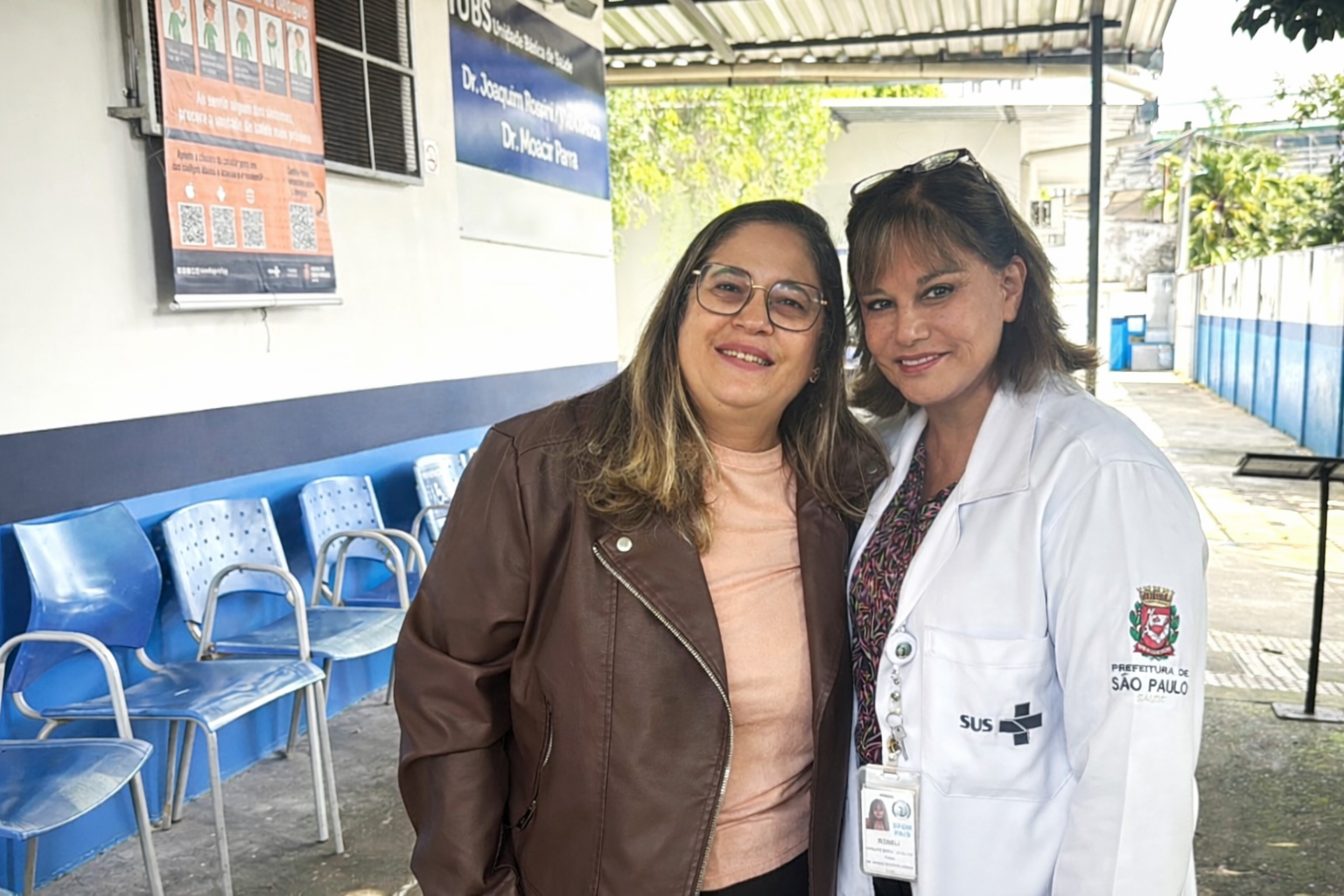 A imagem mostra Simone Vigilato, participante assídua do Avança Saúde vestindo uma blusa em tom claro e uma jaqueta marrom. Ela sorri de forma acolhedora, com postura próxima e confiante, demonstrando vínculo com o serviço.  Ao seu lado, está a gerente da UBS Joaquim Rossini, usando jaleco branco com identificação da Prefeitura de São Paulo e do SUS. Ela também sorri para a câmera, transmitindo segurança e profissionalismo.