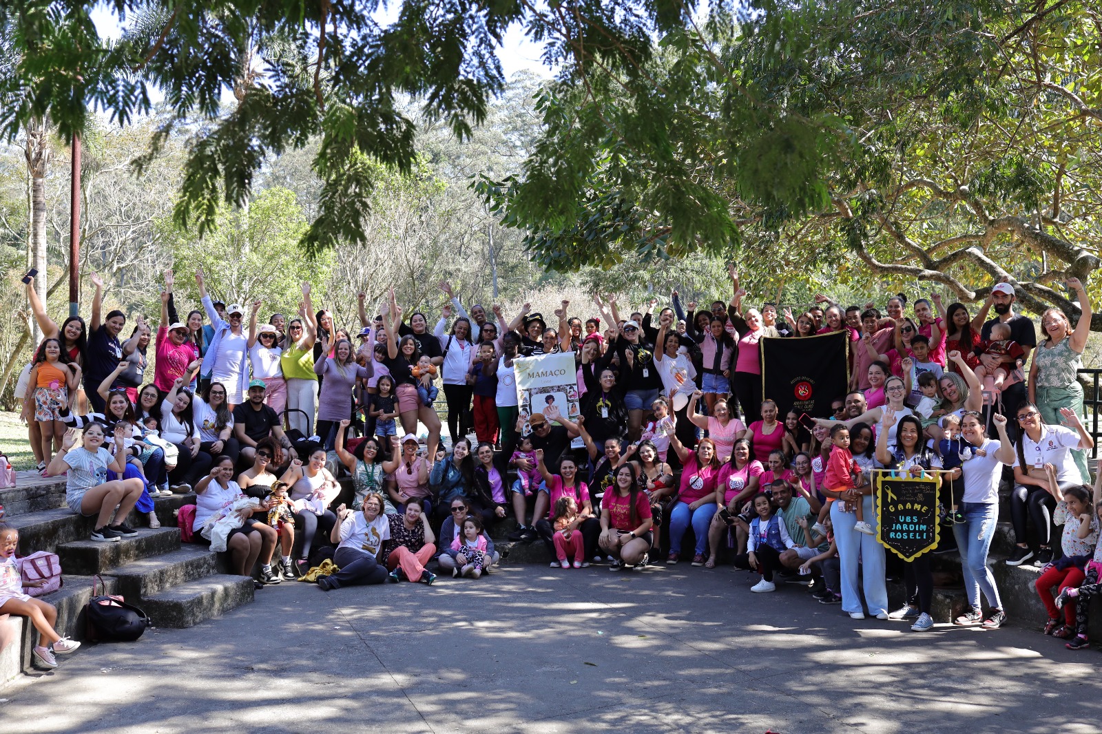 A imagem mostra um grande grupo de pessoas reunidas ao ar livre, em um ambiente arborizado e ensolarado, possivelmente um parque. A maioria está sorrindo, com os braços levantados, celebrando ou posando para a foto em clima de alegria e união. Entre os presentes, há muitas mulheres, crianças, bebês e alguns homens, com destaque para camisetas cor-de-rosa — cor símbolo da amamentação — e cartazes com mensagens relacionadas ao evento.  No centro da imagem, uma faixa com a palavra “Mamaço” indica que se trata de uma ação coletiva de incentivo à amamentação. No canto inferior direito, outro cartaz identifica a participação da UBS Jardim Roseli. As pessoas estão distribuídas em degraus e no chão, em frente a uma área sombreada por árvores, compondo um retrato vibrante de engajamento comunitário.  A imagem expressa acolhimento, diversidade e apoio mútuo, alinhando-se à campanha do Agosto Dourado e à promoção do aleitamento materno.