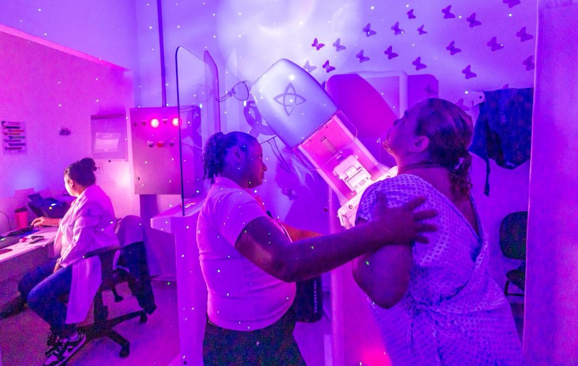 A imagem mostra uma sala de exames médicos iluminada por uma luz arroxeada. À direita, uma paciente veste um avental hospitalar e está posicionada em um equipamento de radiologia — provavelmente para realizar uma mamografia. Uma profissional de saúde a auxilia, ajustando sua postura no aparelho. À esquerda, outra profissional, também de jaleco, está sentada diante de um computador, aparentemente operando o sistema do exame. O ambiente tem uma decoração diferenciada, com projeções de borboletas, estrelas e um luar nas paredes e no teto, criando um clima mais acolhedor e humanizado para o atendimento.