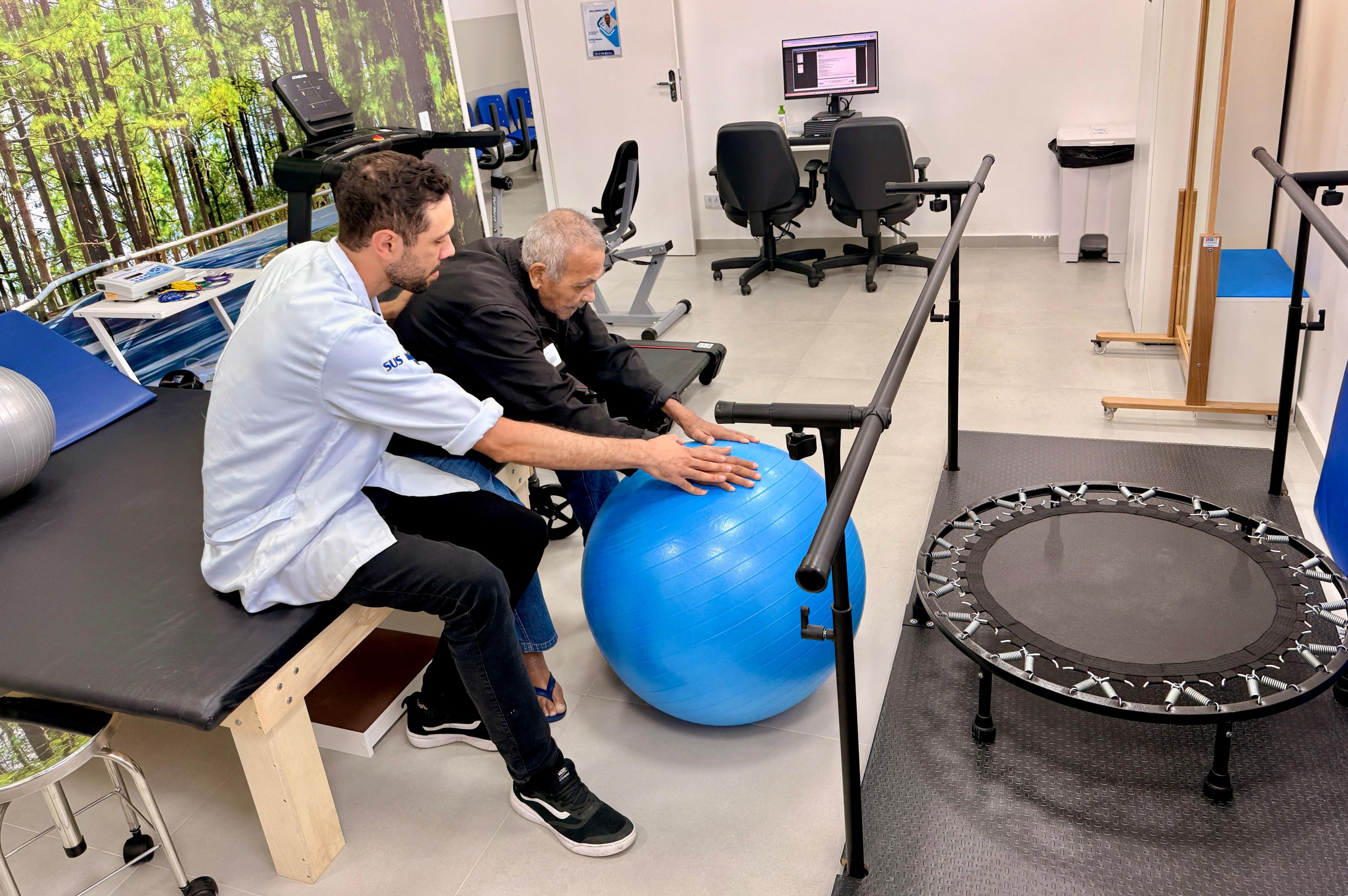A imagem mostra uma sessão de fisioterapia realizada em um ambiente clínico de reabilitação.  Em primeiro plano, um fisioterapeuta vestido com jaleco claro, com a sigla SUS no braço, acompanha um homem idoso sentado ao lado dele. O profissional orienta o paciente durante um exercício com uma bola terapêutica azul (bola suíça), apoiando as mãos sobre ela enquanto o idoso se inclina levemente para frente, indicando um exercício de fortalecimento ou equilíbrio.  O espaço parece ser uma sala de fisioterapia ou reabilitação, equipada com diversos aparelhos. À direita, há barras paralelas para treino de marcha e um mini-trampolim de reabilitação. Ao fundo, é possível ver uma esteira ergométrica, equipamentos de exercício e um computador em uma mesa de atendimento com duas cadeiras.  O ambiente é claro, organizado e bem iluminado, com paredes brancas e uma grande imagem decorativa de uma paisagem de floresta ao fundo, o que ajuda a tornar o espaço mais acolhedor.