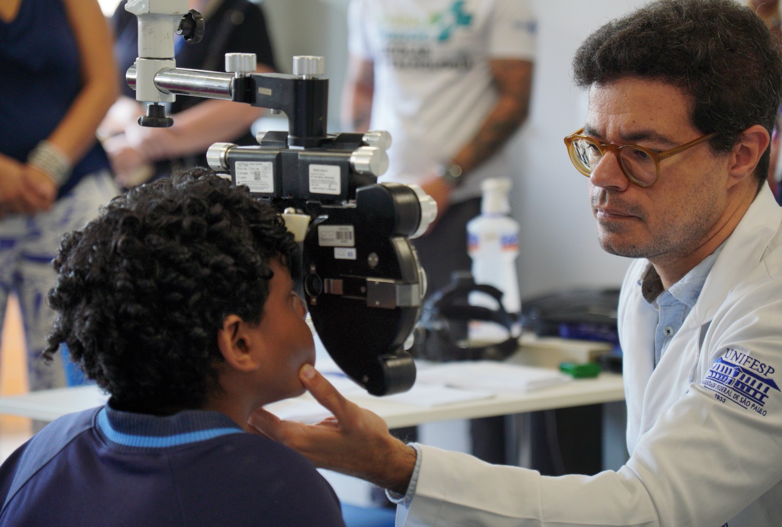 A imagem mostra um atendimento oftalmológico sendo realizado em uma criança. O paciente, um menino, está sentado diante de um equipamento de exame de visão (provavelmente um autorrefrator ou parte de um exame oftalmológico), enquanto um médico o observa atentamente. O profissional de saúde, usando jaleco branco e óculos, posiciona delicadamente a cabeça da criança com a mão, garantindo o alinhamento correto para o exame. Ele demonstra concentração e cuidado durante o procedimento. No jaleco, é possível ver o logotipo de uma instituição de ensino/saúde. Ao fundo, há outras pessoas desfocadas, sugerindo um ambiente clínico ou uma ação de atendimento coletivo, como mutirão ou triagem. Sobre a mesa, aparecem itens típicos de consultório, como frascos de álcool ou produtos de higienização.