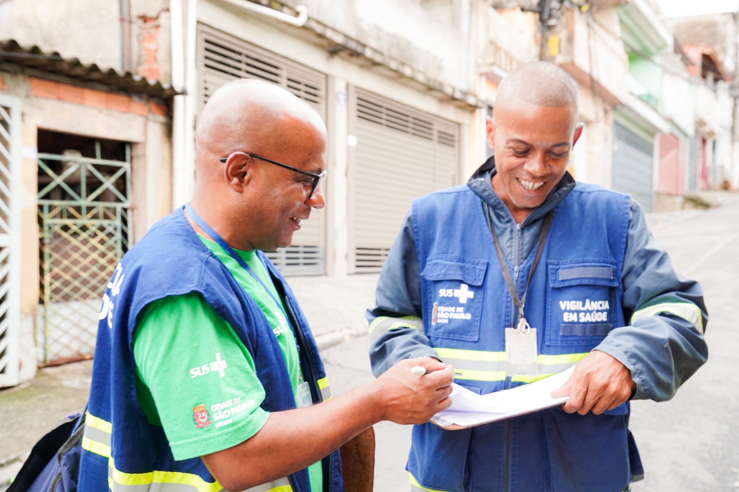 A imagem mostra dois homens negros sorrindo enquanto conversam e fazem anotações em uma prancheta, em uma rua residencial. Eles usam coletes azuis com faixas refletivas e o emblema do SUS, além da inscrição “Vigilância em Saúde” e “Cidade de São Paulo – Saúde”. Um deles veste camiseta verde por baixo do colete e óculos; o outro está de jaqueta azul e segura uma caneta. O cenário é uma rua estreita, com casas simples e portões metálicos, típicos de um bairro urbano.
