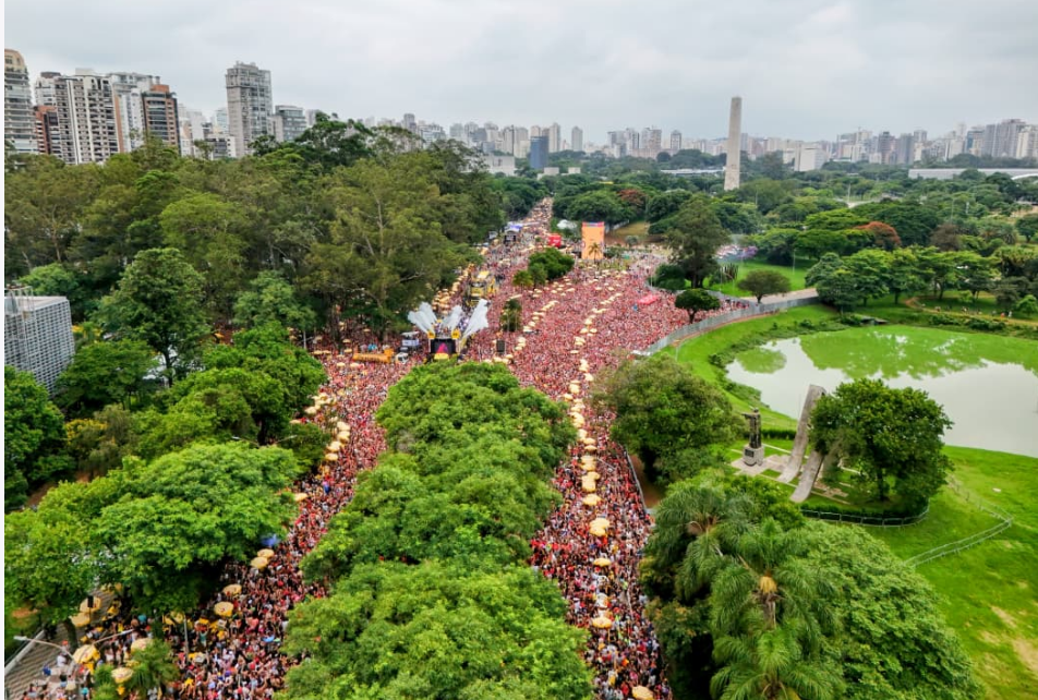 A imagem retrata a abertura do Carnaval de rua na cidade de São Paulo, com uma multidão impressionante reunida no Parque do Ibirapuera. Vista de cima, a avenida que corta o parque aparece completamente tomada por foliões, formando um grande corredor humano que se estende por entre as árvores e áreas verdes.  No centro do percurso, destaca-se a estrutura do trio elétrico, onde acontece o show de Ivete Sangalo, responsável por conduzir a festa e embalar o público com seus sucessos. A concentração de pessoas evidencia a dimensão do evento, considerado o maior Carnaval de rua do país, reunindo milhares de pessoas em clima de celebração.  Ao redor, o verde do parque, o lago e os monumentos contrastam com a energia vibrante da multidão, enquanto ao fundo surgem os prédios da cidade, reforçando a ideia de São Paulo como palco dessa grande festa popular. A cena transmite alegria, diversidade e a força do carnaval paulistano abrindo oficialmente a folia na capital