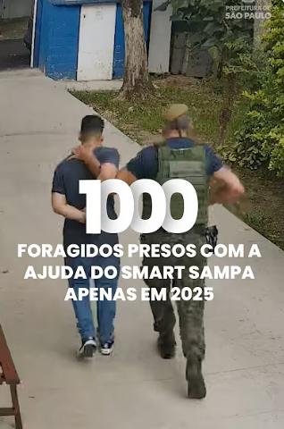 A imagem mostra uma cena de detenção ao ar livre. Um homem, com uma camiseta azul escura, calças jeans e tênis, está sendo escoltado por um agente de segurança. O agente, de costas para o observador, veste um uniforme camuflado e um colete tático escuro, e parece estar algemando ou segurando o braço do homem detido por trás.  Em primeiro plano, sobreposta à cena, há um grande número branco "1000" e o texto em português, também em branco: "FORAGIDOS PRESOS COM A AJUDA DO SMART SAMPA APENAS EM 2025".