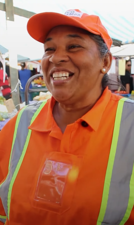 Na imagem, uma pessoa está usando um uniforme de cor laranja com detalhes refletivos amarelos e prateados. Ela veste uma camisa tipo polo com gola e um colete de segurança por cima. Também está de boné laranja. No fundo, é possível ver um ambiente aberto, que parece ser uma feira ou mercado ao ar livre, com tendas e outras pessoas ao redor.
