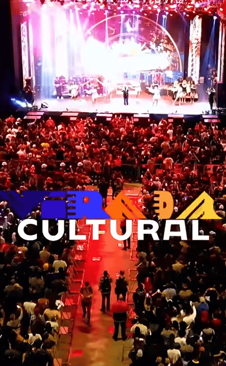 iluminado por luzes coloridas, há músicos e artistas se apresentando diante de uma plateia numerosa. O público ocupa quase todos os assentos, e algumas pessoas caminham pelo corredor central. Sobre a imagem, em destaque, está o texto "VIRADA CULTURAL", com "VIRADA" em letras estilizadas nas cores azul, laranja e amarela, e "CULTURAL" em branco. A atmosfera é vibrante e festiva, destacando a celebração da cultura.