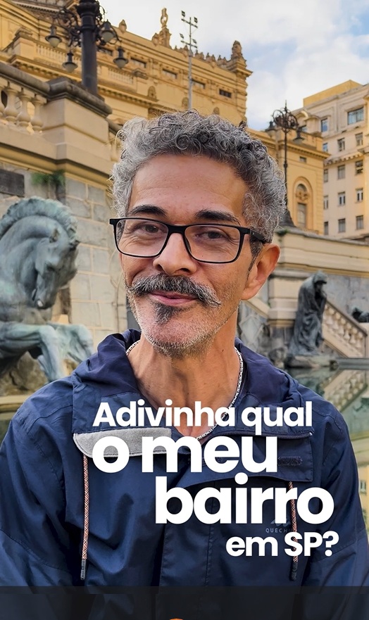 Homem de cabelos grisalhos e barba curta veste uma jaqueta azul e está posicionado em frente ao Theatro Municipal de São Paulo, um edifício histórico com detalhes arquitetônicos clássicos e esculturas de cavalos ao fundo. No centro da imagem, aparece o texto: "Adivinha qual o meu bairro em SP?" em letras brancas. O céu está parcialmente nublado e há prédios antigos ao redor.