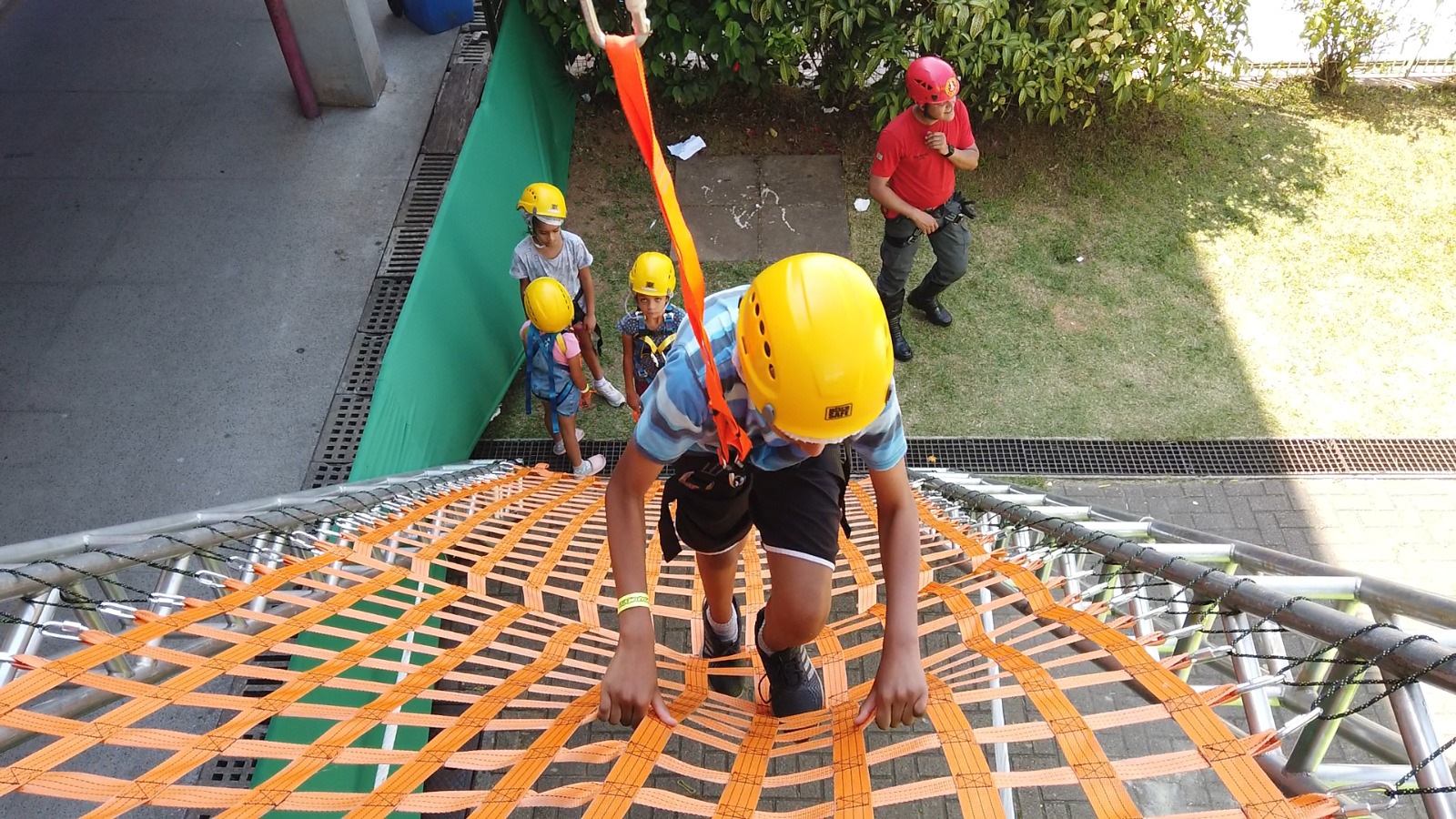A imagem mostra um grupo de crianças participando de uma atividade de escalada em uma estrutura de cordas, sob supervisão de um adulto. Todas as crianças estão usando capacetes de segurança amarelos e cintos de proteção com fitas, subindo uma rede de fitas laranjas presa a uma armação metálica. Uma das crianças está em destaque, quase no topo da estrutura, enquanto as outras aguardam na base, junto ao instrutor que veste camisa vermelha e capacete próprio. O ambiente ao redor possui uma área gramada, plantas e uma calçada, indicando que a atividade ocorre em um espaço aberto e seguro.