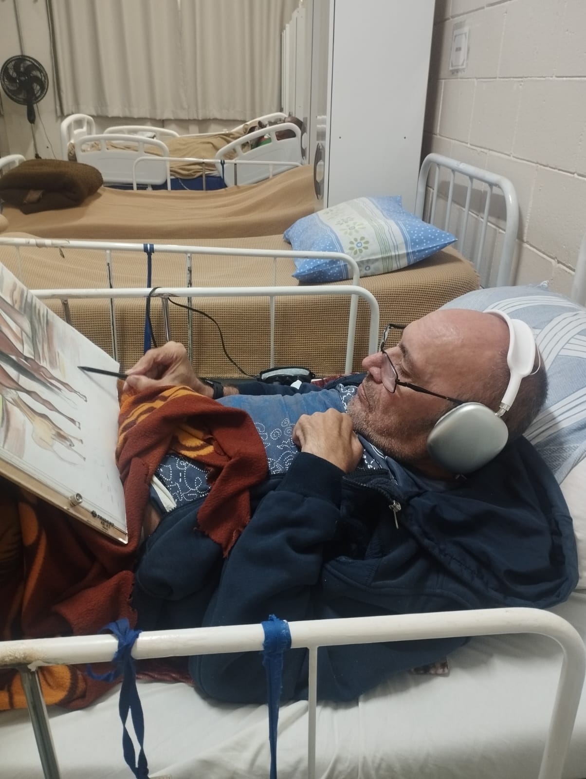 Senhor residente de serviço de ILPI está deitado numa cama leito fazendo atividade de pintura para exposição.