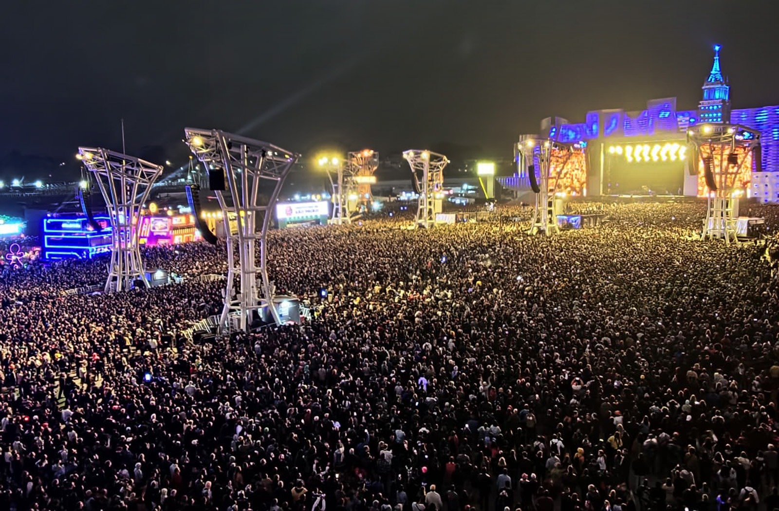 A imagem mostra uma multidão em um festival de música noturno ao ar livre. O palco principal é uma estrutura elaborada que se assemelha a uma cidade iluminada por luzes de neon, com prédios de várias alturas e cores.