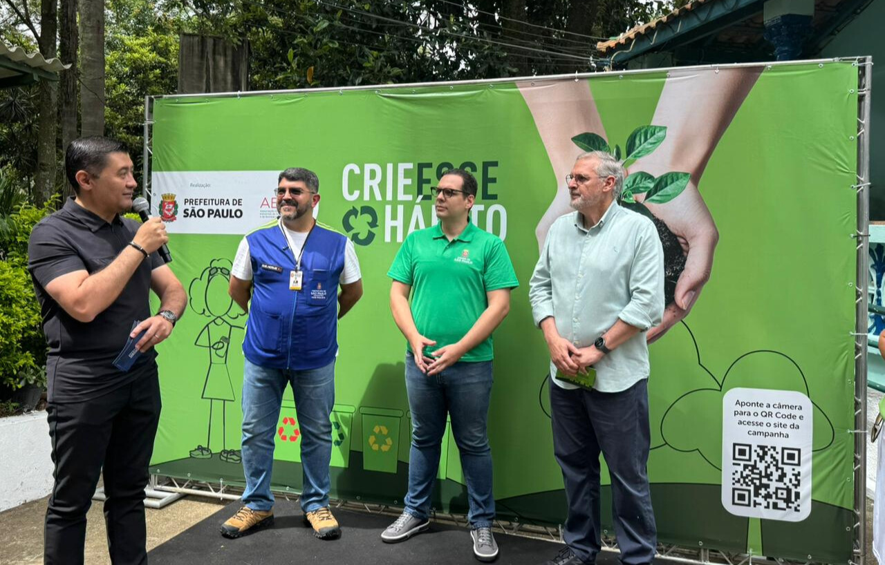 Em área externa, quatro homens estão diante de um painel verde da campanha “Crie Esse Hábito”. À esquerda, um homem fala ao microfone enquanto os outros observam. O painel mostra símbolos de reciclagem, uma muda de planta e um QR Code, indicando ação de conscientização ambiental.