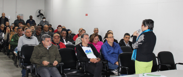 A imagem mostra uma sala de reuniões ou auditório com dezenas de pessoas sentadas, assistindo atentamente a uma mulher que fala ao microfone. A palestrante está de pé, de costas parcialmente voltadas para a câmera, e usa calça verde-limão, blusa preta com detalhes brancos nas mangas e um cachecol azul. O público é composto majoritariamente por adultos e idosos, homens e mulheres, vestindo roupas casuais de frio. Algumas pessoas usam crachás e há cadeiras pretas organizadas em fileiras. O ambiente é simples, com paredes brancas e iluminação artificial. Ao fundo, um ventilador de parede está ligado e algumas pessoas permanecem em pé. A cena sugere uma reunião comunitária ou capacitação, com foco em participação cidadã ou assuntos públicos.