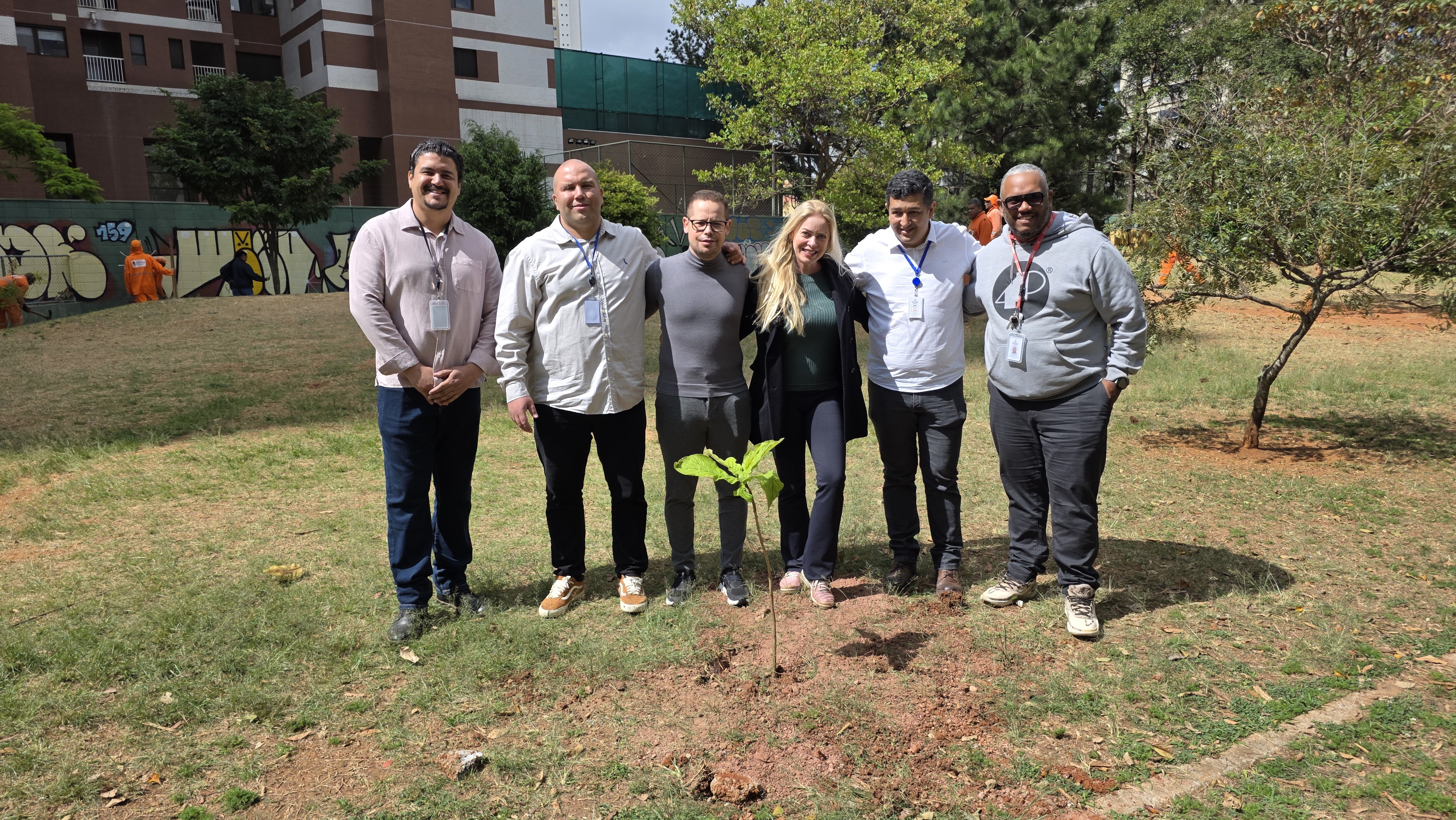 Imagem 1: Grupo de pessoas, três homens ao lado esquerdo, uma mulher de cabelos loiros ao centro e mais dois homens ao lado direito, reunidos em área verde após o plantio de uma muda de árvore. Imagem 2: Dois homens, um de camisa cinza e outro de camisa branca realizando o plantio de uma muda de árvore em área verde.