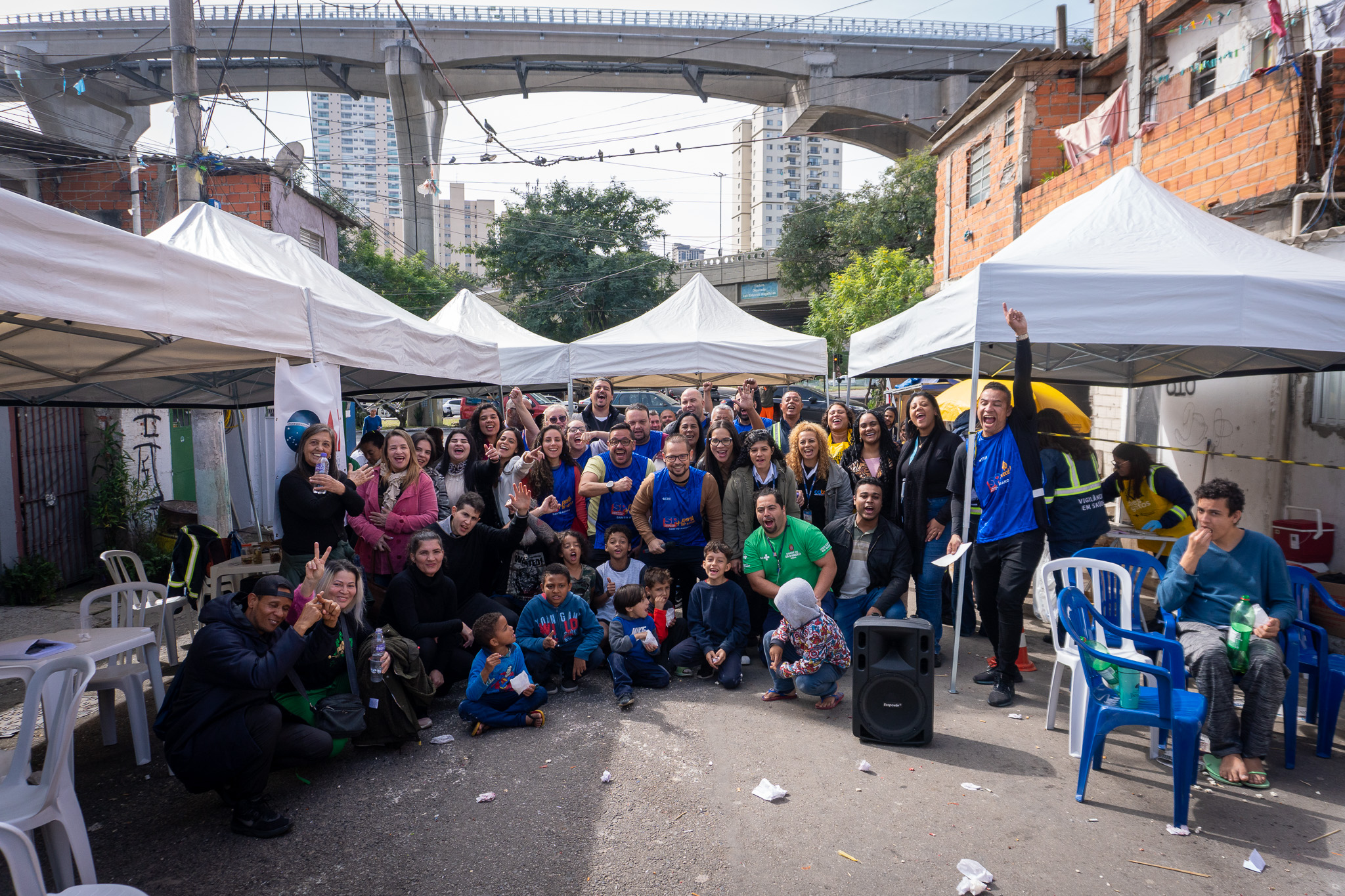 Créditos da foto: Kennedy Silva Descritivo da imagem: Foto de um grupo grande de pessoas sorrindo e fazendo poses de alegria no meio de uma rua com tendas brancas montadas. Há adultos e crianças, algumas sentadas e outras em pé, com prédios e um viaduto ao fundo. Foto de um grupo grande de pessoas sorrindo e fazendo poses de alegria no meio de uma rua com tendas brancas montadas. Há adultos e crianças, algumas sentadas e outras em pé, com prédios e um viaduto ao fundo.