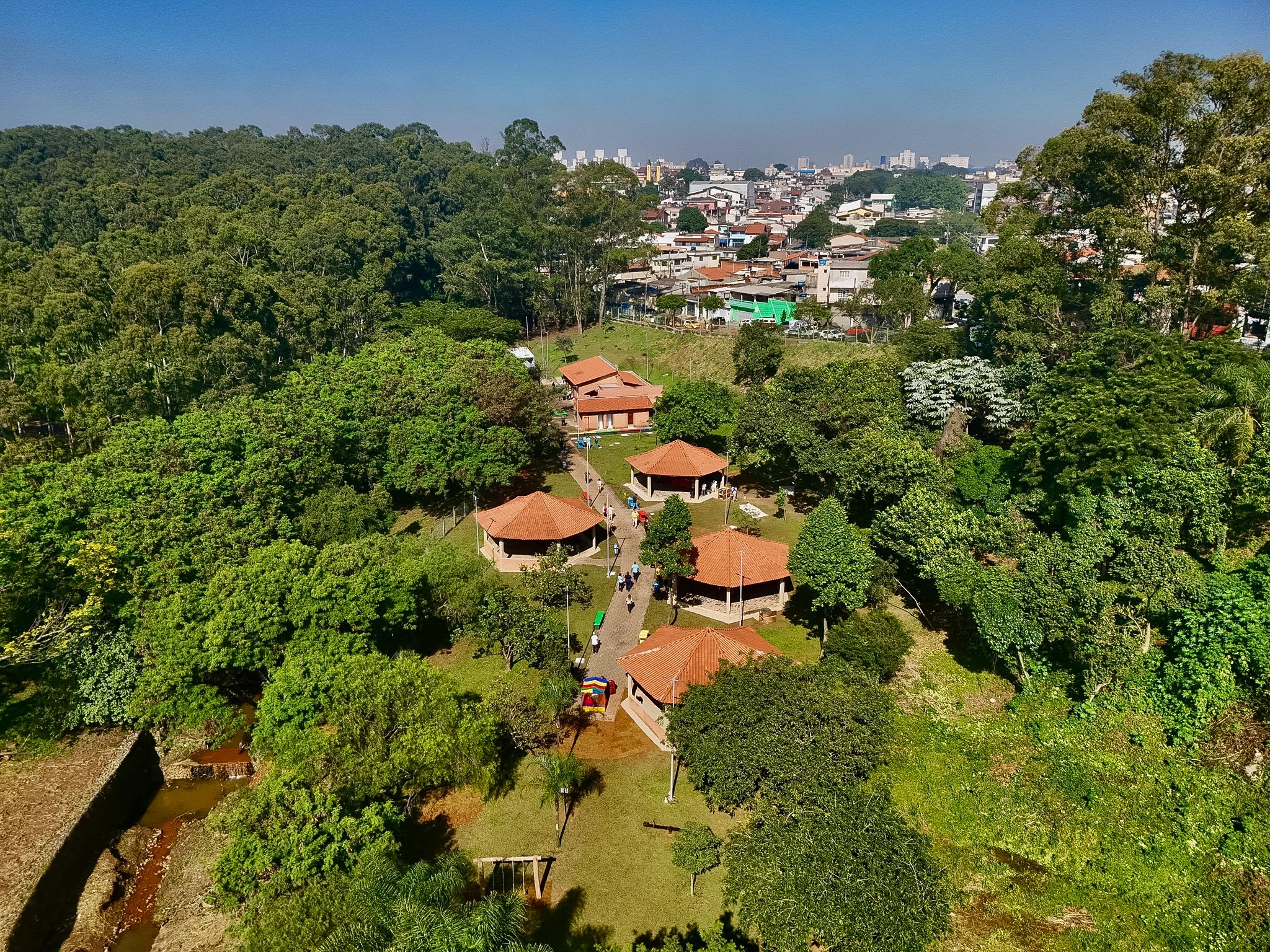 Fotografia aérea que mostra a área de lazer ao ar livre do parque, em um dia ensolarado. O local possui diversas construções com telhados de barro e pessoas circulando por caminhos pavimentados.