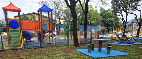 A imagem mostra o trecho do caneteiro central da Av. Inajar de Souza revistalizado. A cena tem luz natural de dia, com céu nublado  e demonstra  cuidado com o espaço comunitário.  Principais elementos:  Parquinho infantil (à esquerda): um conjunto de brinquedos multicoloridos — laranja, azul, amarelo e verde — com torres cobertas por telhados piramidais (laranja e azul). Inclui um escorregador azul, escadas e proteções laterais. A área é cercada por grades metálicas coloridas (verdes, amarelas e vermelhas) e o piso é de placas emborrachadas quadradas com cores alternadas.  Academia ao ar livre (à direita): equipamentos de ginástica de metal, pintados principalmente de azul e amarelo.  Área de convivência (centro, em primeiro plano): uma mesa redonda de concreto sobre uma base pintada de azul, acompanhada de quatro banquinhos circulares isolados, pintados de verde.  Ambiente e fundo: há áreas de grama pelo terreno. Ao fundo, Composição: é um plano geral que reúne os três espaços de uso — brincar, se exercitar e conviver — em um mesmo ambiente, mostrando como a praça foi pensada para atender diferentes públicos e atividades.