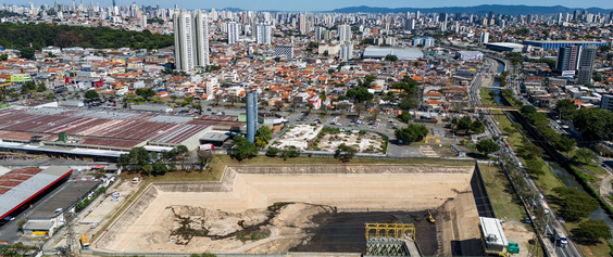 A imagem é uma vista aérea, fotografada de cima para baixo, de uma grande área urbana densamente povoada, provavelmente em um dia ensolarado, com luz forte e clara que sugere um período próximo ao meio-dia.  🔵 A cena principal  O elemento central é uma grande bacia de contenção de águas pluviais — uma escavação retangular e profunda, como um 