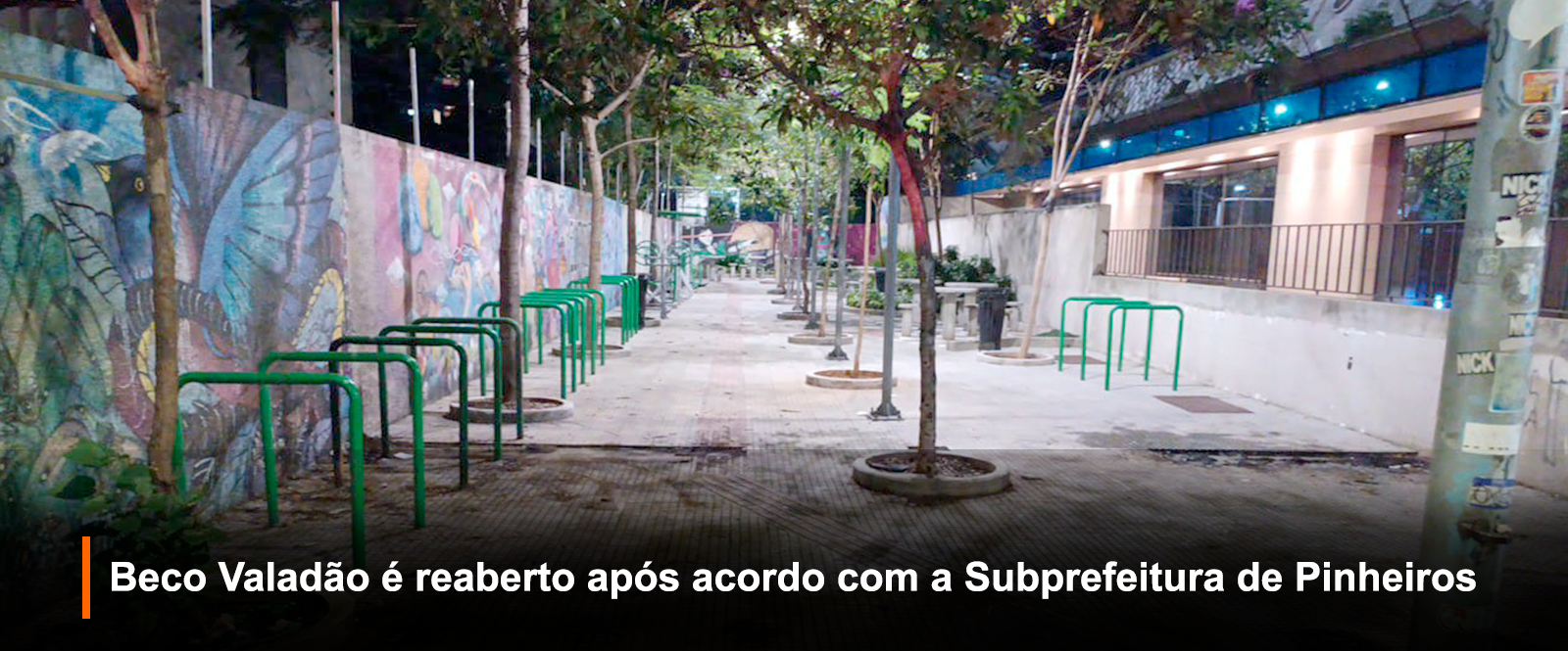 Fotografia noturna em ambiente urbano mostra o Beco Valadão em Pinheiros, São Paulo, iluminado. O local tem piso de ladrilhos, árvores e bicicletários. Um mural colorido com grafites ocupa a esquerda, enquanto um prédio moderno com janelas azuis aparece à direita.  Uma faixa preta translúcida na parte inferior anuncia em destaque: 