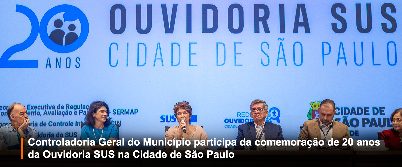 Fotografia de evento formal em auditório: seis pessoas em mesa de autoridades. Painel azul ao fundo com a logomarca dos 20 anos da Ouvidoria SUS da Cidade de São Paulo. Faixa preta à frente: 
