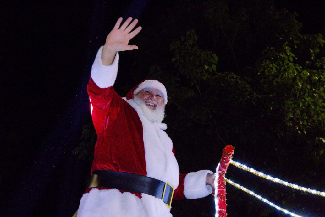 #Pracegover: Close-up noturno, de cintura para cima, do Papai Noel. Ele está alegremente sorrindo e acenando com a mão direita levantada, vestindo seu tradicional traje vermelho e branco com um cinto preto largo, barba e cabelos brancos volumosos. Ele parece estar em um carro alegórico ou palco iluminado, segurando uma barra decorada com luzes pisca-pisca, com um fundo predominantemente escuro, onde se pode notar levemente a folhagem de árvores no topo.