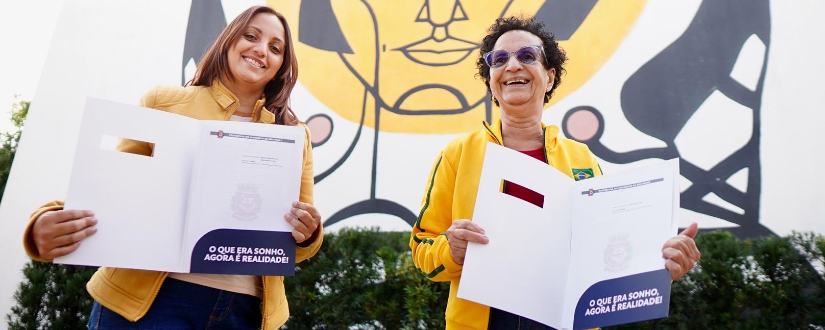 Duas mulheres felizes segurando suas novas escrituras de suas casas, dentro das pastas a fresa: 
