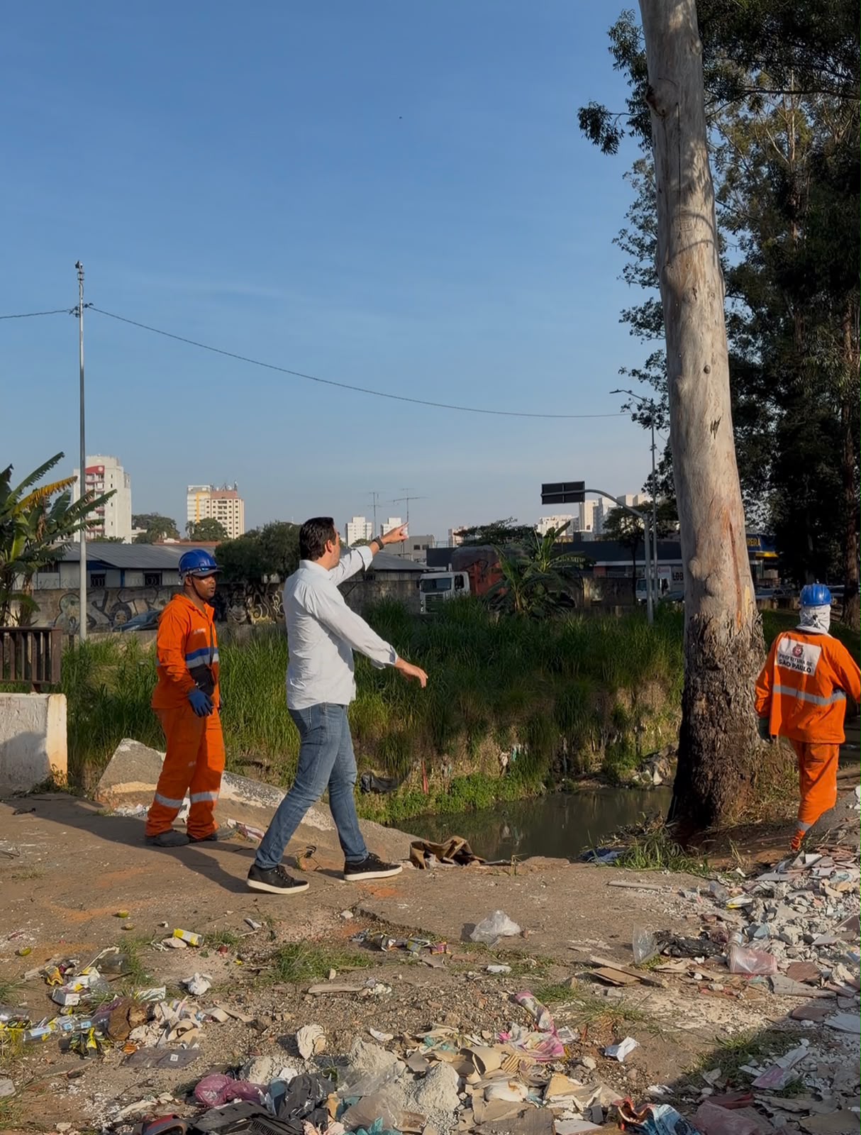 Foto em plano médio e ângulo normal, tirada ao ar livre durante o dia, sob luz solar direta.  No centro da imagem, um homem de pele clara, cabelos escuros e calça jeans, usando camisa social clara de mangas compridas dobradas até o antebraço, caminha em direção à direita enquanto aponta com o braço direito estendido para frente.  À esquerda, um trabalhador veste uniforme laranja de sinalização com faixas refletivas, capacete azul e luvas, acompanhando o movimento do homem. À direita, de costas para a câmera, outro trabalhador com o mesmo uniforme laranja e capacete azul caminha perto do tronco de uma grande árvore alta, de casca clara. Na parte posterior do uniforme desse trabalhador, lê-se a inscrição "PREFEITURA DE SÃO PAULO".  O chão no primeiro plano está repleto de entulho, pedaços de concreto e lixo espalhado. Ao fundo, vê-se um canal ou córrego com vegetação em suas margens, além de edifícios urbanos e vegetação sob um céu azul limpo.