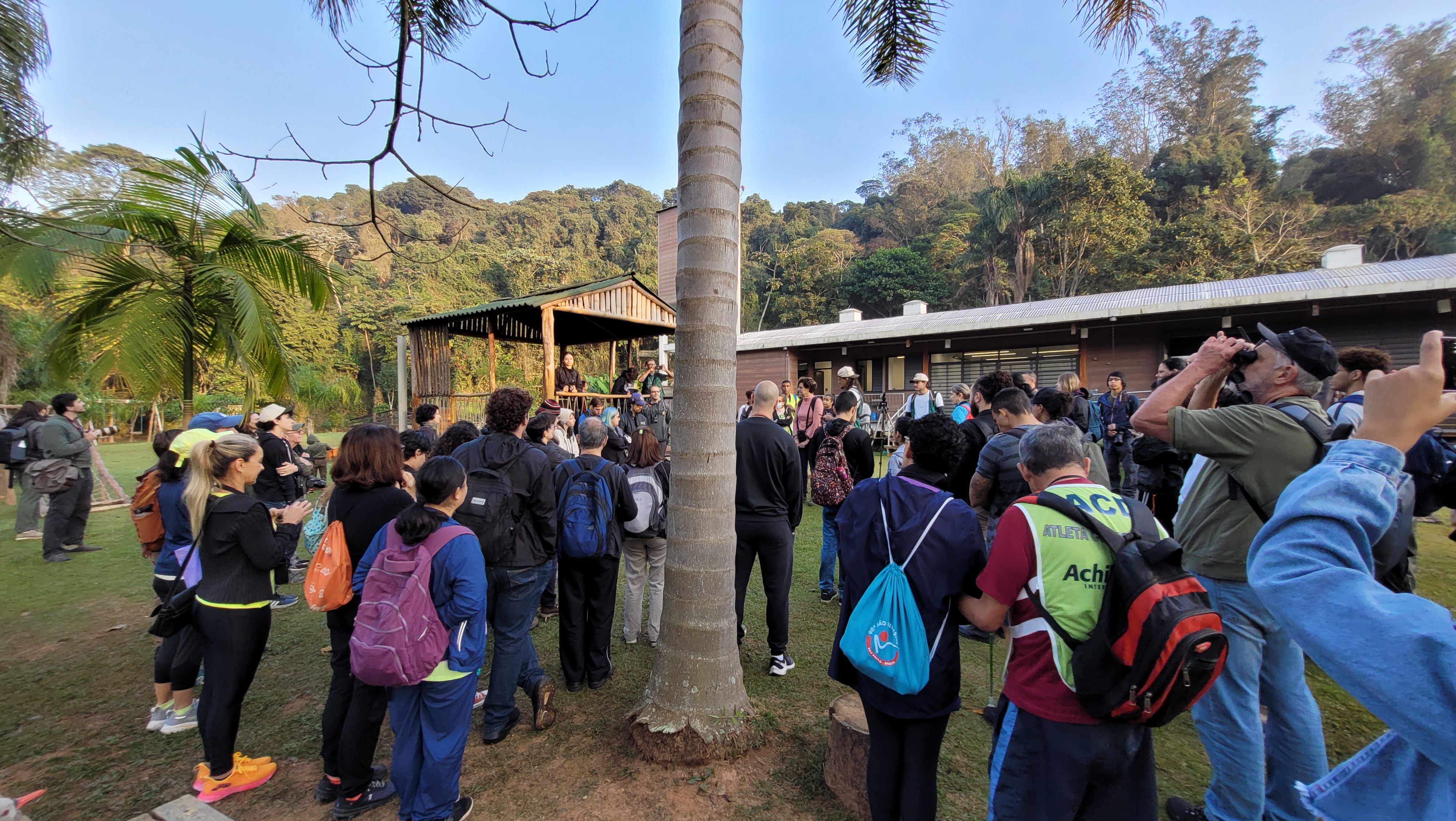 Grupo de pessoas reunidas em área gramada com árvores e construções de madeira ao fundo. Algumas pessoas estão de mochila, outras usam binóculos para observar o ambiente. O encontro acontece em espaço natural cercado de mata.