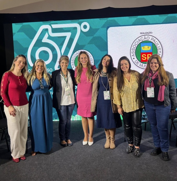 Sete mulheres estão lado a lado em um palco, sorrindo, durante o 67º Congresso da Associação Paulista de Municípios. Com estilos diversos e crachás no pescoço, elas representam a presença feminina em espaços de liderança e diálogo público. Ao fundo, um telão com o logotipo do evento.