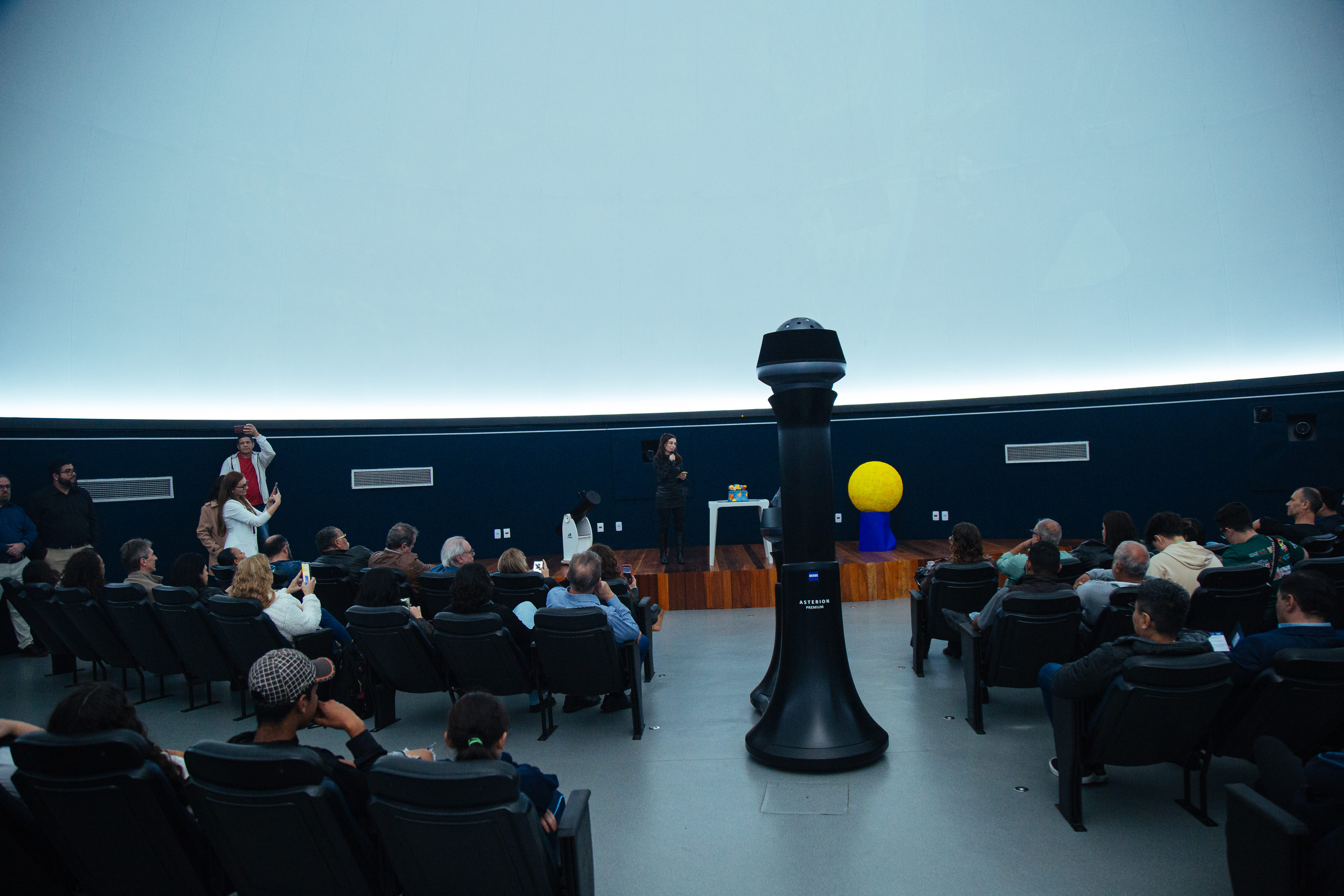 A imagem mostra o interior do Planetário de Parelheiros, com várias pessoas sentadas em poltronas, assistindo a uma apresentação. Uma mulher está em pé no palco, falando ao microfone. O ambiente é escuro, com iluminação suave na base da cúpula, típica de planetários. Ao fundo, há objetos decorativos relacionados ao espaço, como um modelo do Sol e telescópios. O público é diverso e atento.
