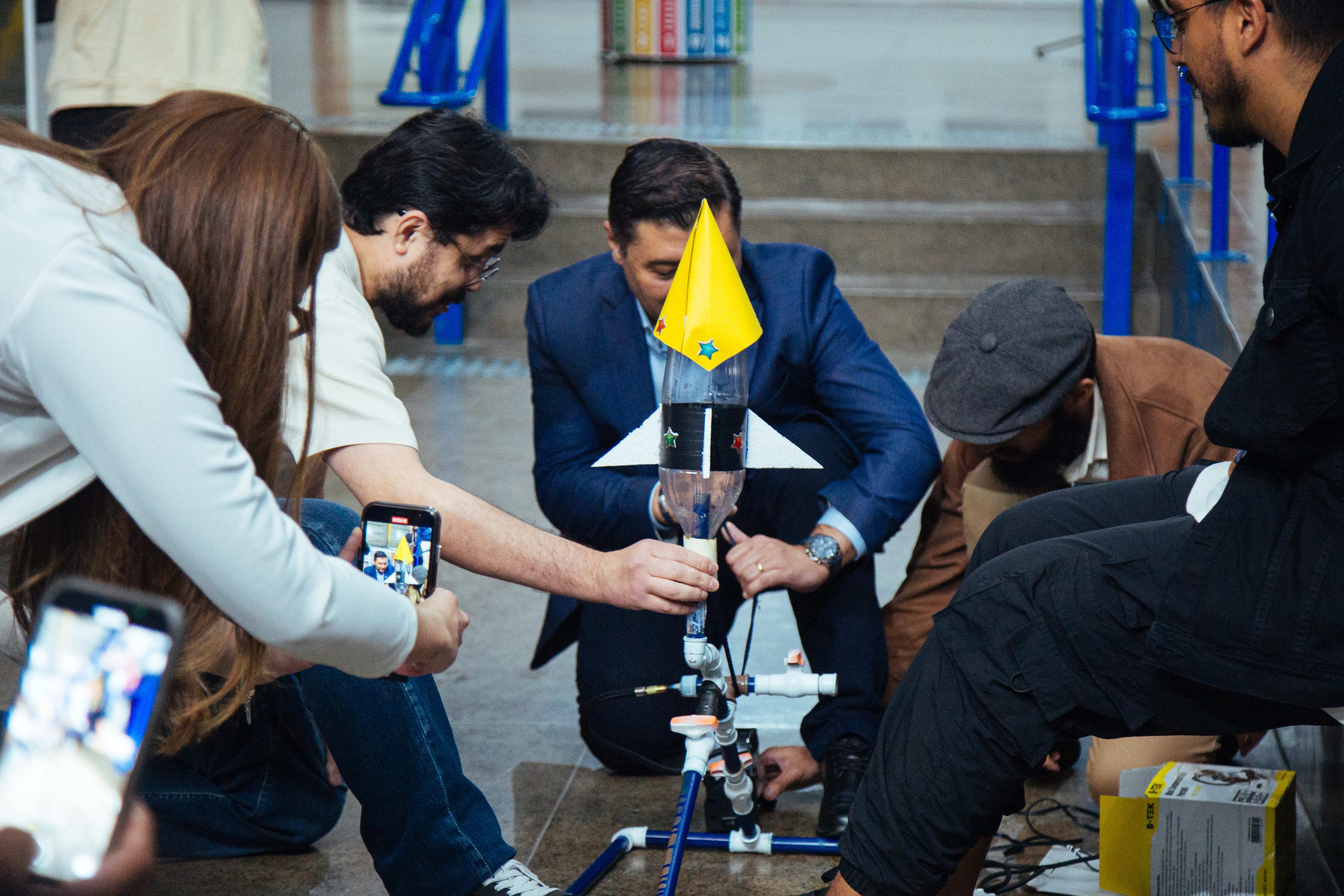 A imagem mostra um grupo diverso de pessoas reunido em um ambiente interno, montando um foguete artesanal feito com uma garrafa plástica. Alguns estão agachados, manuseando o foguete e a base de lançamento, enquanto outros registram o momento com celulares. O clima é de colaboração e entusiasmo.