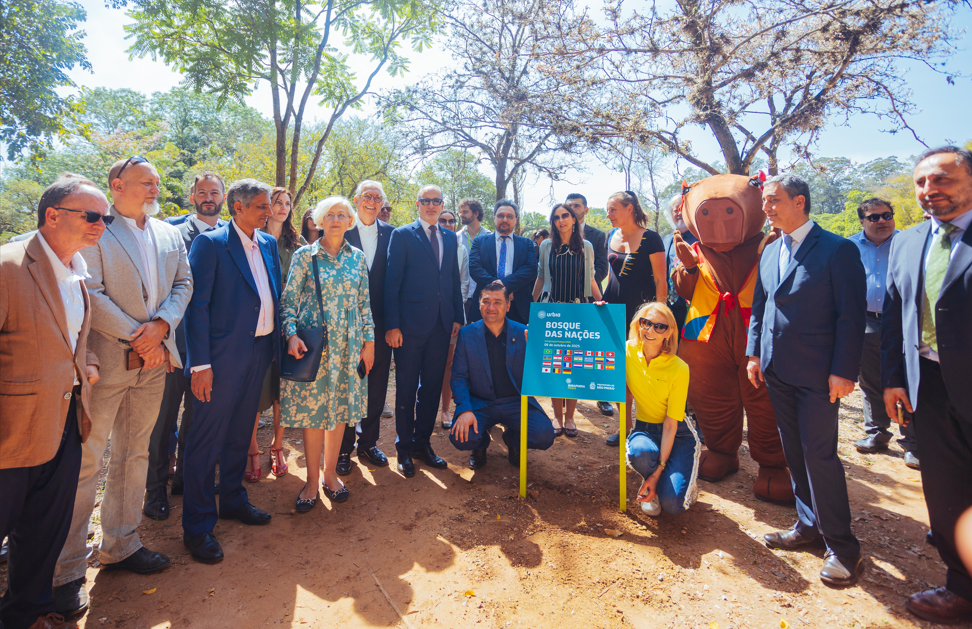 Na imagem, um grupo de autoridades e representantes consulares participa da inauguração do Bosque das Nações, no Parque Ibirapuera, em São Paulo. No centro, há uma placa azul com o nome do projeto e as bandeiras dos países envolvidos. O evento simboliza a cooperação internacional e a valorização da diversidade cultural por meio do plantio de espécies nativas de diferentes nações.  Entre os presentes, estão diplomatas, representantes da Prefeitura de São Paulo e da Secretaria do Verde e do Meio Ambiente (SVMA), além da mascote em formato de capivara, que traz um toque simbólico e descontraído à cerimônia.
