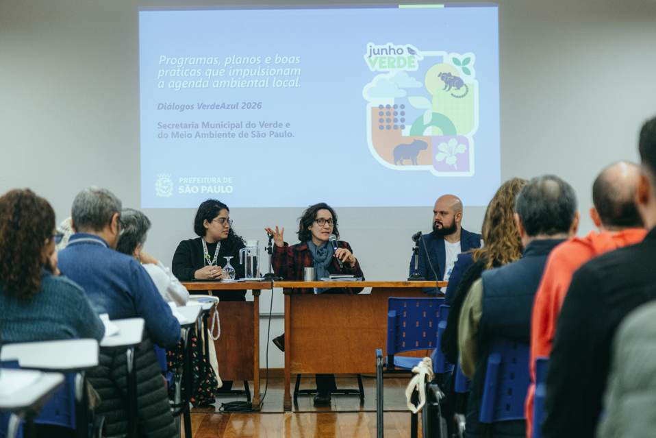 Fotografia de um seminário realizado em auditório. Ao centro, três pessoas estão sentadas atrás de uma mesa participando de um painel de debates. A mulher no centro fala ao microfone enquanto gesticula com uma das mãos. Ao fundo, uma tela de projeção exibe uma apresentação do programa Junho Verde, da Secretaria Municipal do Verde e do Meio Ambiente de São Paulo, com o tema “Programas, planos e boas práticas que impulsionam a agenda ambiental local”. Na plateia, diversas pessoas acompanham a discussão sentadas em fileiras voltadas para os palestrantes. O ambiente é fechado, iluminado e voltado à troca de conhecimentos sobre temas ambientais.