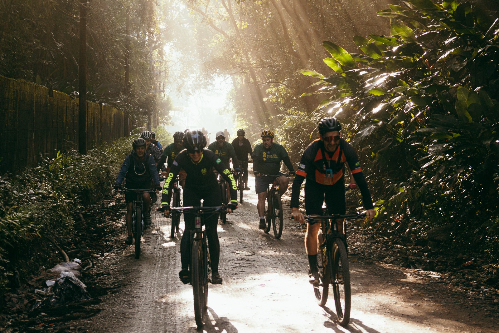 Fotografia de um grupo de ciclistas pedalando por uma estrada de terra cercada por vegetação densa. Os participantes usam capacetes e roupas esportivas, enquanto os raios de sol atravessam as copas das árvores e iluminam o caminho.