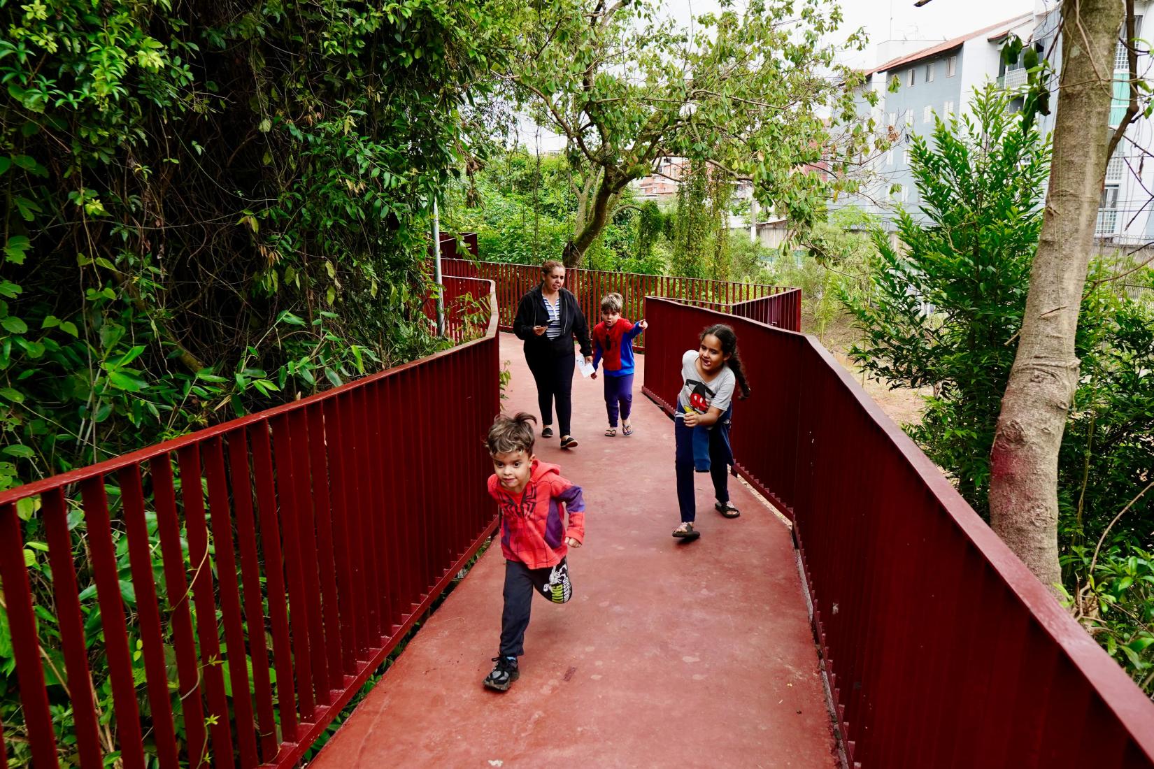 Imagem em um parque com bastante vegetação. Em primeiro plano, uma passarela com piso avermelhado e corrimãos laterais também vermelhos segue em leve curva. À esquerda e à direita, há árvores e plantas densas, com folhas verdes de diferentes tamanhos.  Quatro pessoas caminham pela passarela. Na frente, uma criança pequena corre em direção à câmera, usando casaco vermelho e calça escura. Logo atrás, uma mulher adulta caminha acompanhando outras duas crianças. Uma delas veste roupa colorida e anda no centro da passarela; a outra, à direita, parece sorrir enquanto caminha, apoiando-se levemente no corrimão.  Ao fundo, entre a vegetação, aparecem edifícios residenciais de poucos andares, indicando que o parque está inserido em uma área urbana. A cena transmite movimento e uso do espaço por famílias.