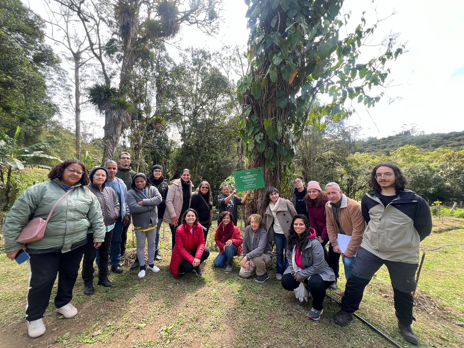 Grupo diverso de cerca de 18 pessoas, em área verde, sorrindo para a foto. Duas pessoas seguram uma placa verde em uma árvore que indica área de preservação permanente. O ambiente transmite união e conexão com a natureza.